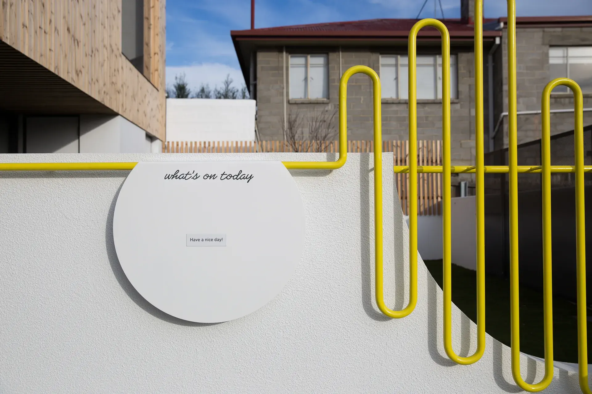 This white wall features a circular information board, a vibrant yellow pipe runs along the top of the wall and transitions into a wavy, slatted fence design.