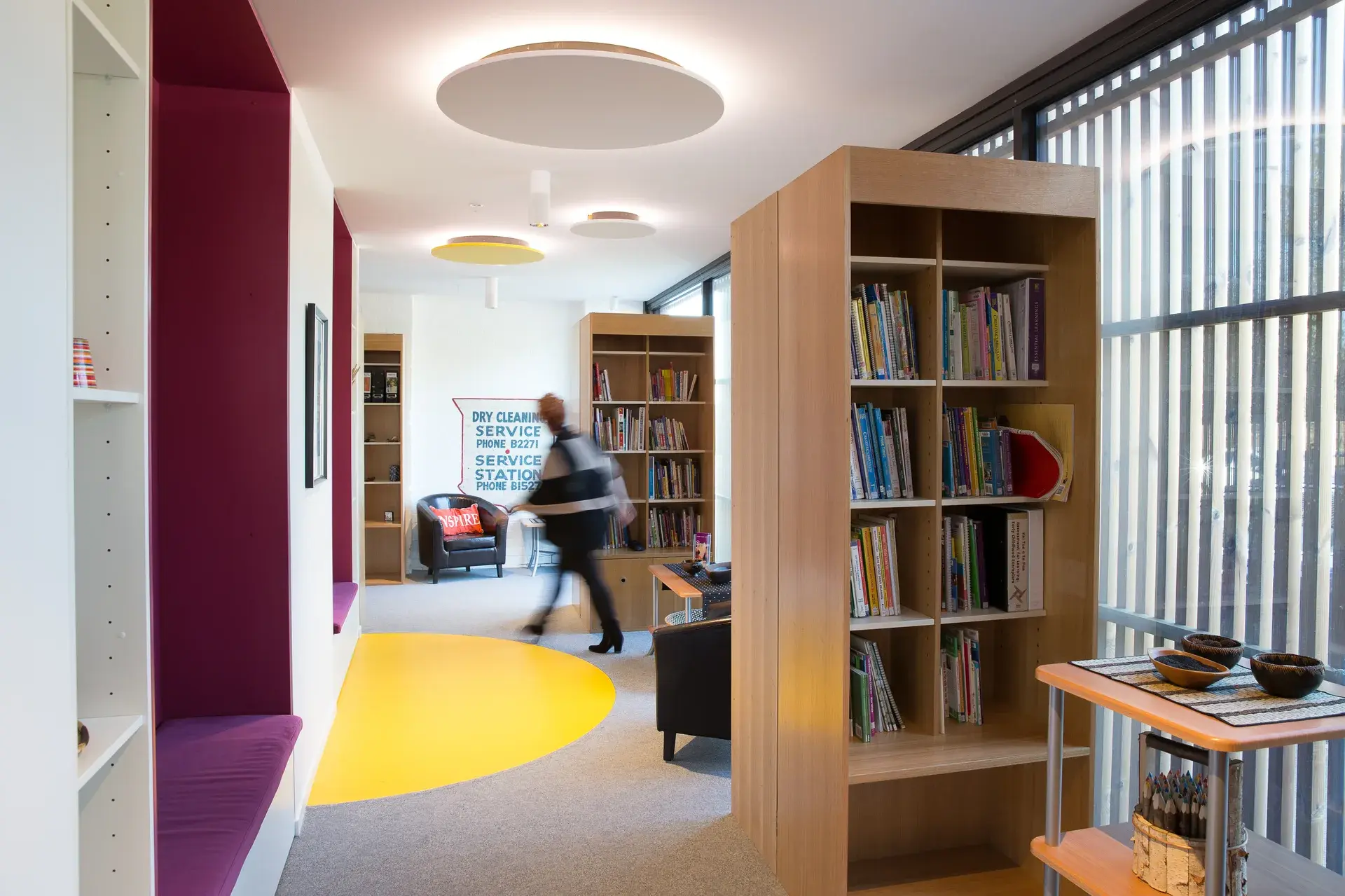 This library or reading area features tall wooden bookshelves and built-in purple cushioned seating recessed into the wall. A vibrant yellow circle is painted on the grey carpeted floor, while slatted wood screens filter natural light from the large windows.