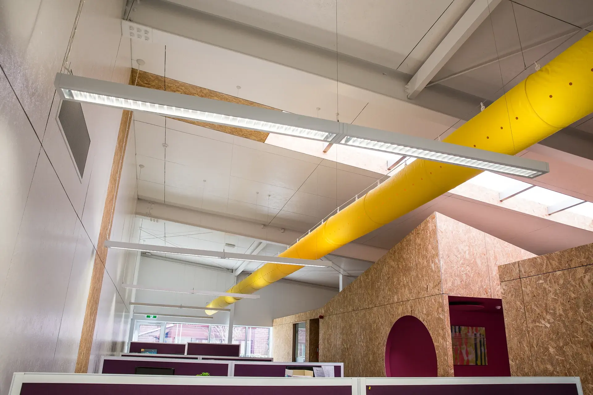 This open-plan office features a large, bright yellow ventilation duct running across the ceiling above purple-topped cubicles. In the background, a structure made of OSB (oriented strand board) contains a recessed pink nook with a circular entrance.
