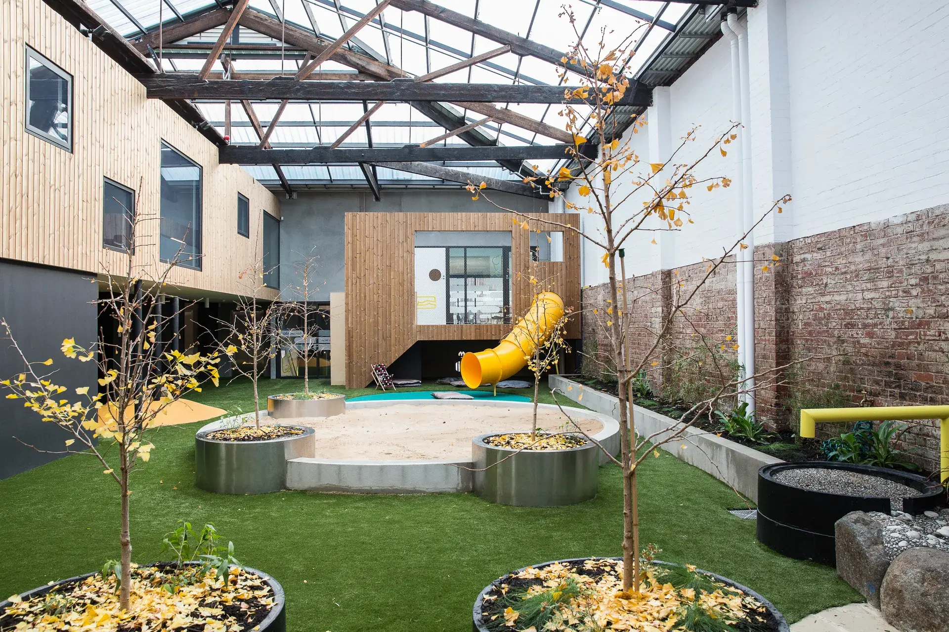 Gemini said This indoor courtyard features a central sandpit, artificial turf, and a bright yellow slide. The space is enclosed by wood-panelled and brick walls under a high, glass-panelled ceiling with industrial timber trusses.