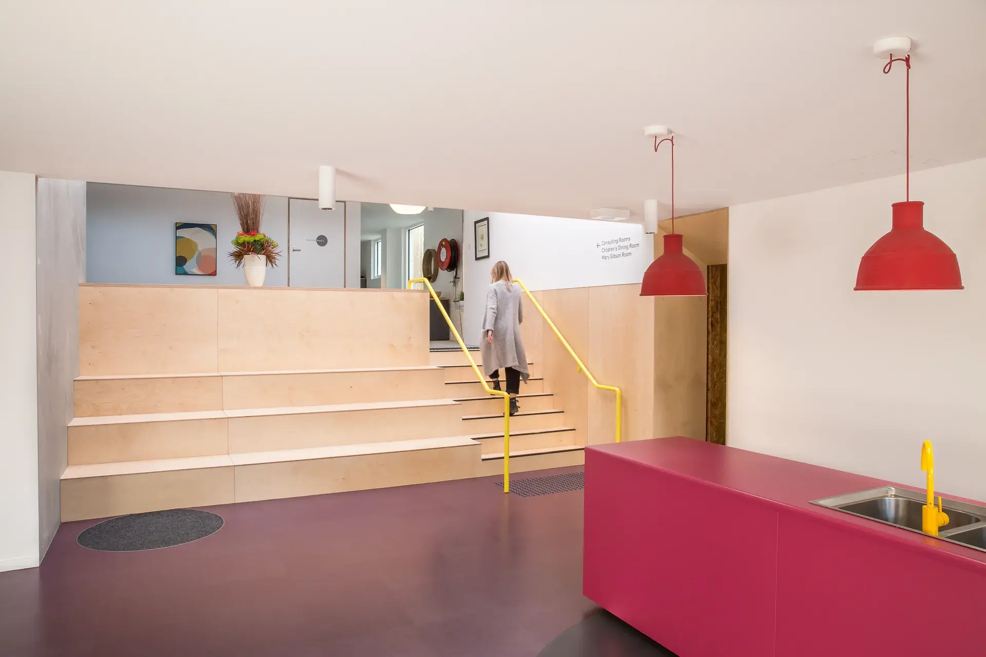 This indoor area features a large wooden tiered seating structure and a vibrant pink counter with a yellow faucet. A person is walking up the stairs, which are lined with vibrant yellow railing. Red pendant lights hang from the ceiling, adding a pop of colour to the neutral white walls.