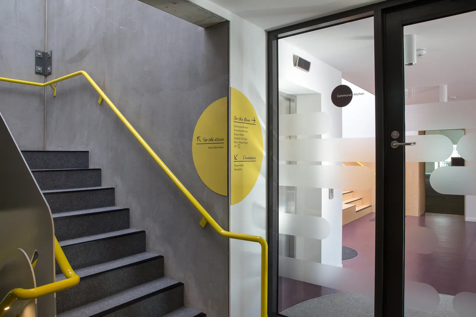 This staircase features dark grey steps and a vibrant yellow railing against a textured concrete wall. A large yellow circular sign provides floor directions next to a glass door.