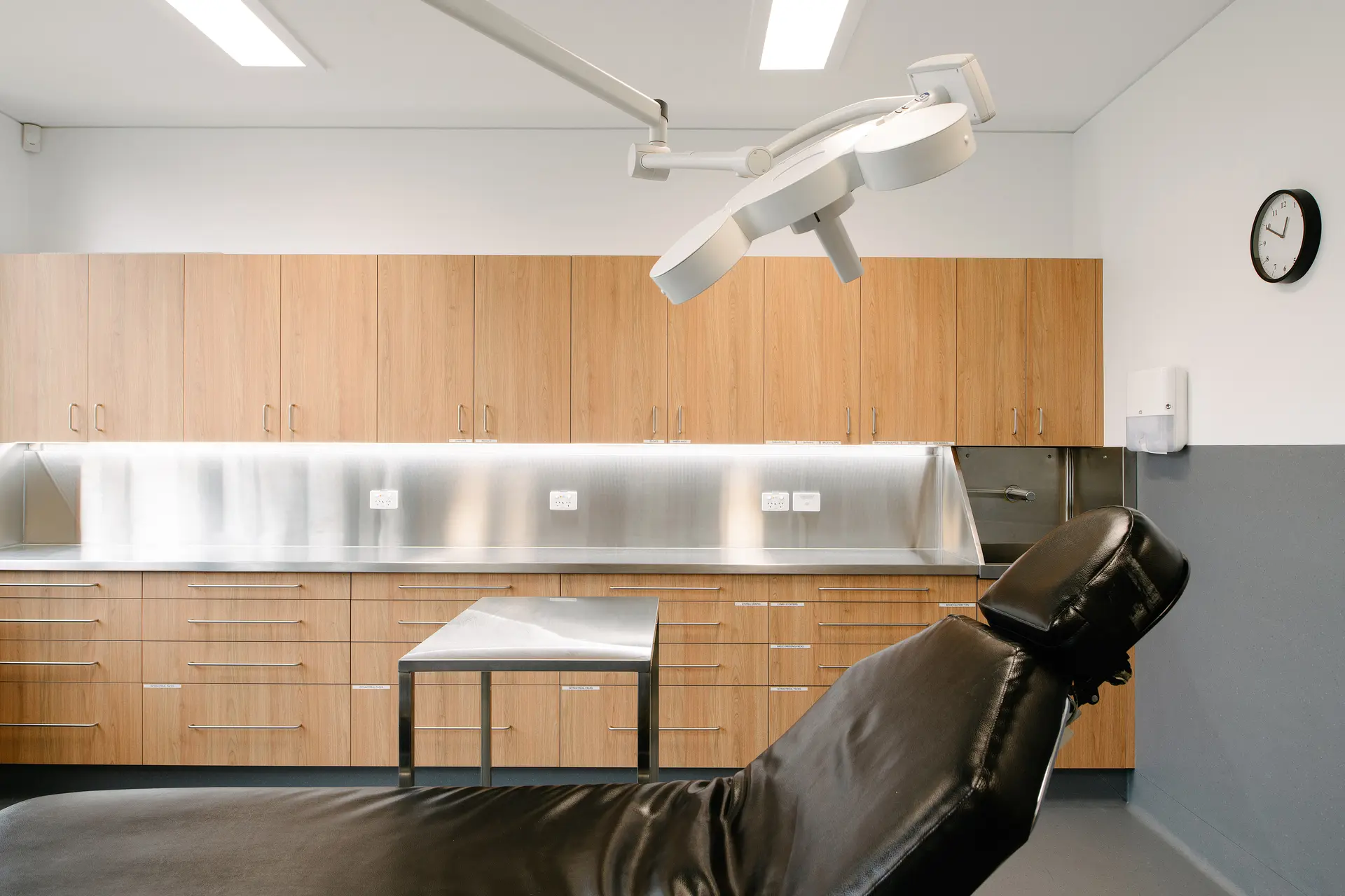 A clean medical procedure room featuring a black examination chair, a surgical light, stainless steel counters, and light wood cabinetry.