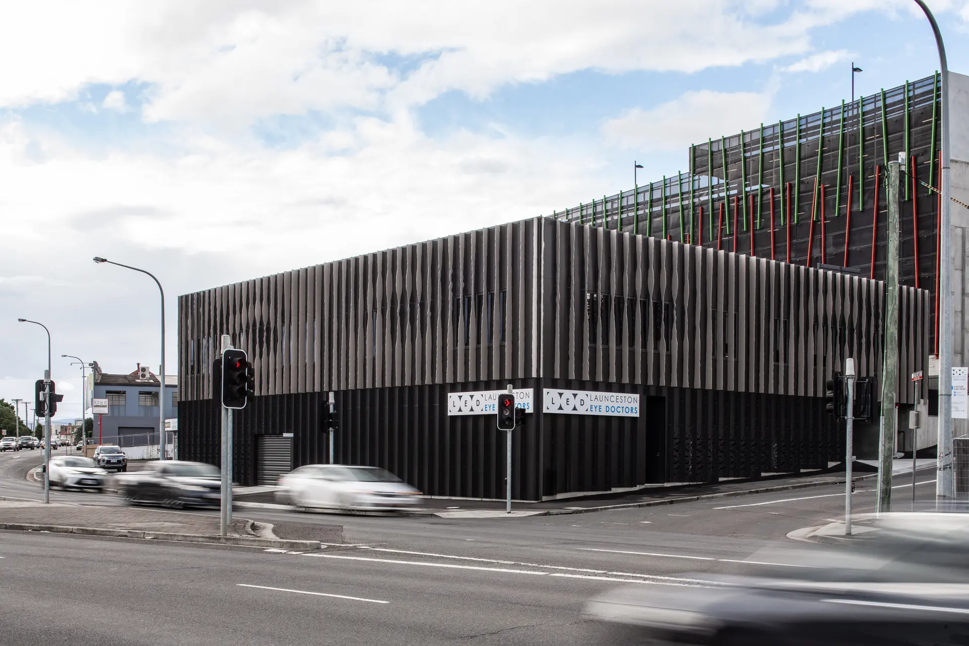A close-up of the façade reveals layered, folded metal panels in light grey tones, their subtle shifts in depth and alignment producing a moiré-like effect that resolves into eye-like forms as one moves past the building.