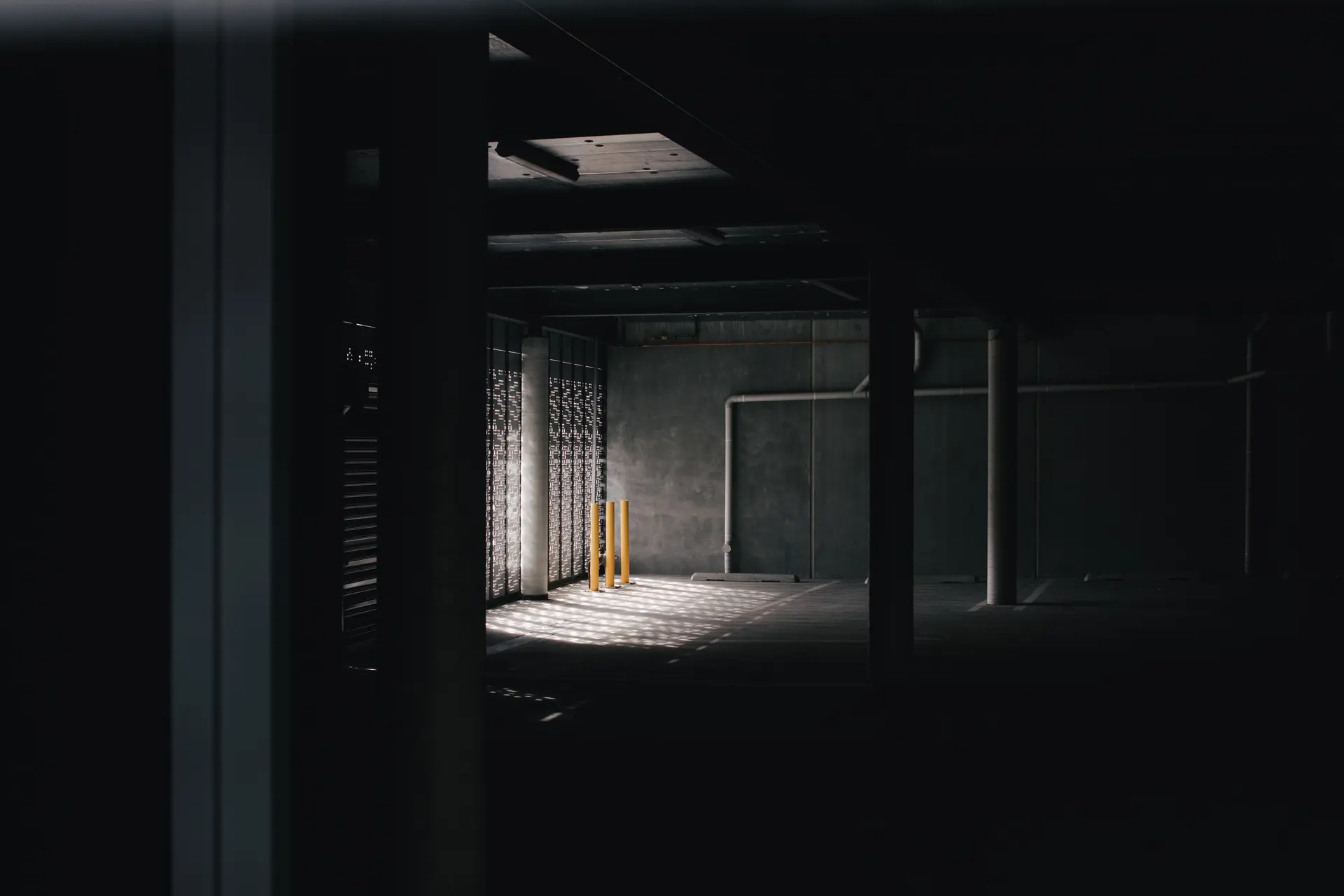 Sunlight filters through a perforated screen into a dark concrete parking garage, illuminating three yellow bollards.