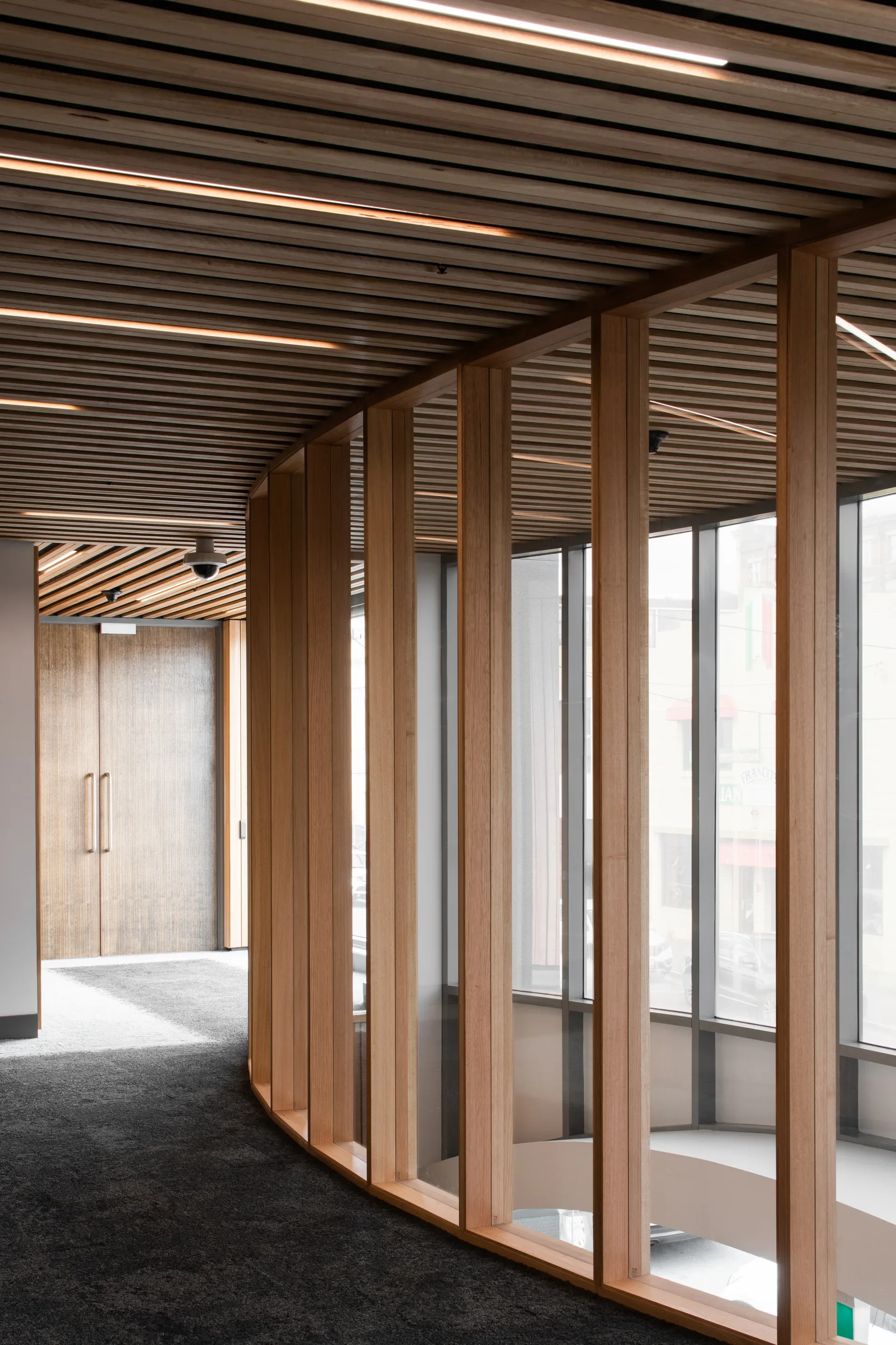 A curved glass wall with vertical timber frames follows a dark carpeted hallway toward a set of wooden double doors. Above, the ceiling features thin wooden slats with integrated linear lighting.