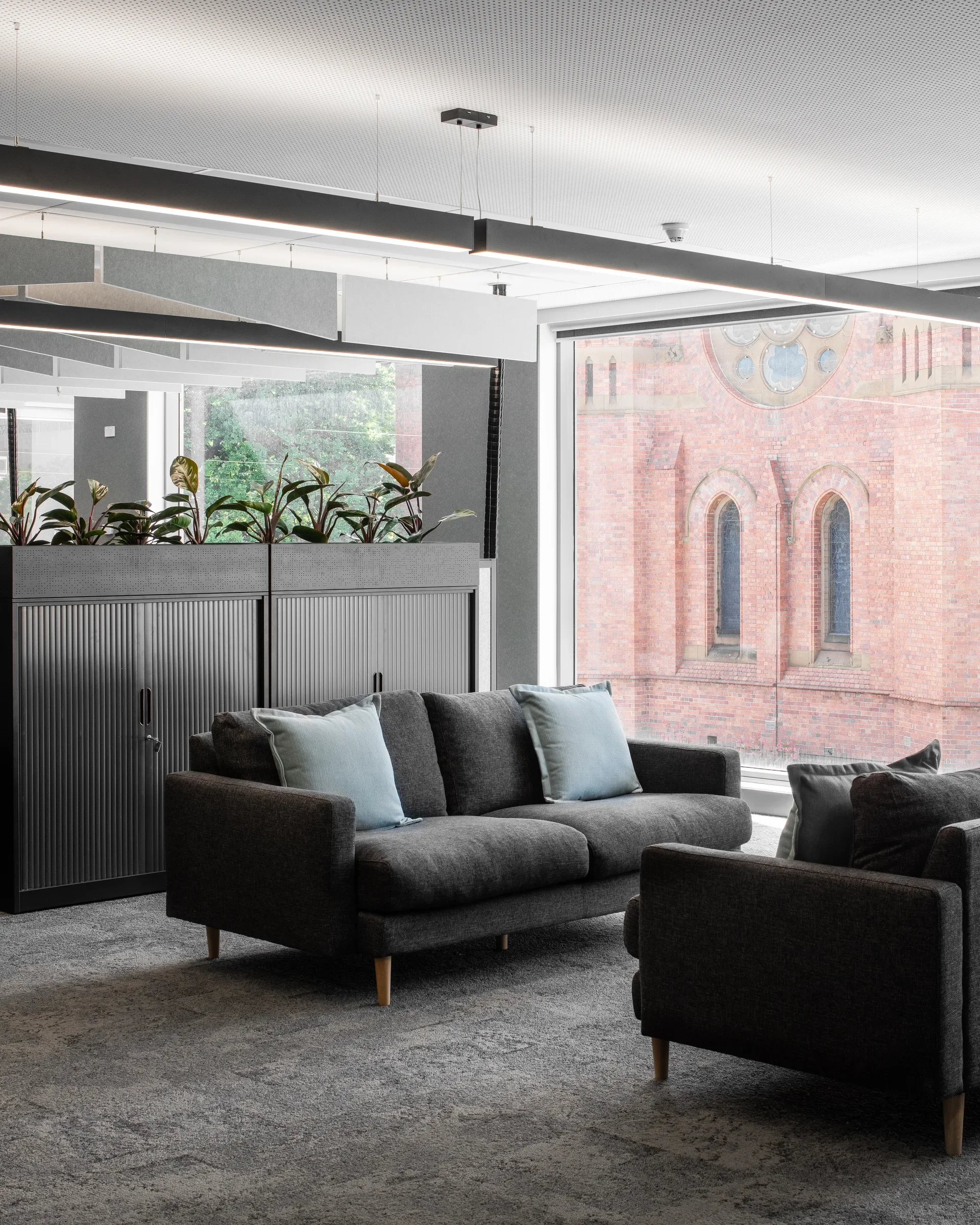 Grey sofas on a speckled carpet face a large window overlooking a historic red-brick building. Indoor plants sit atop low cabinets beneath modern linear lighting.