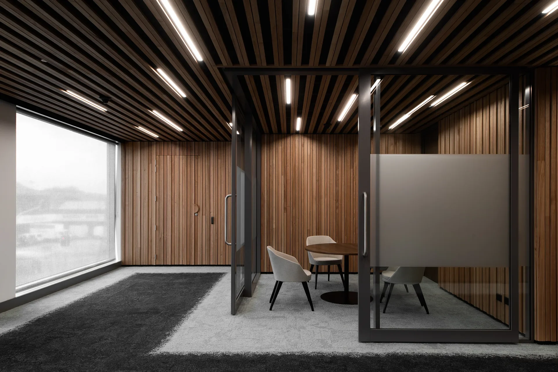 Three cream chairs sit around a circular table in a meeting room enclosed by glass with a frosted privacy strip. The space features vertical timber walls and a matching slatted ceiling with integrated lighting.