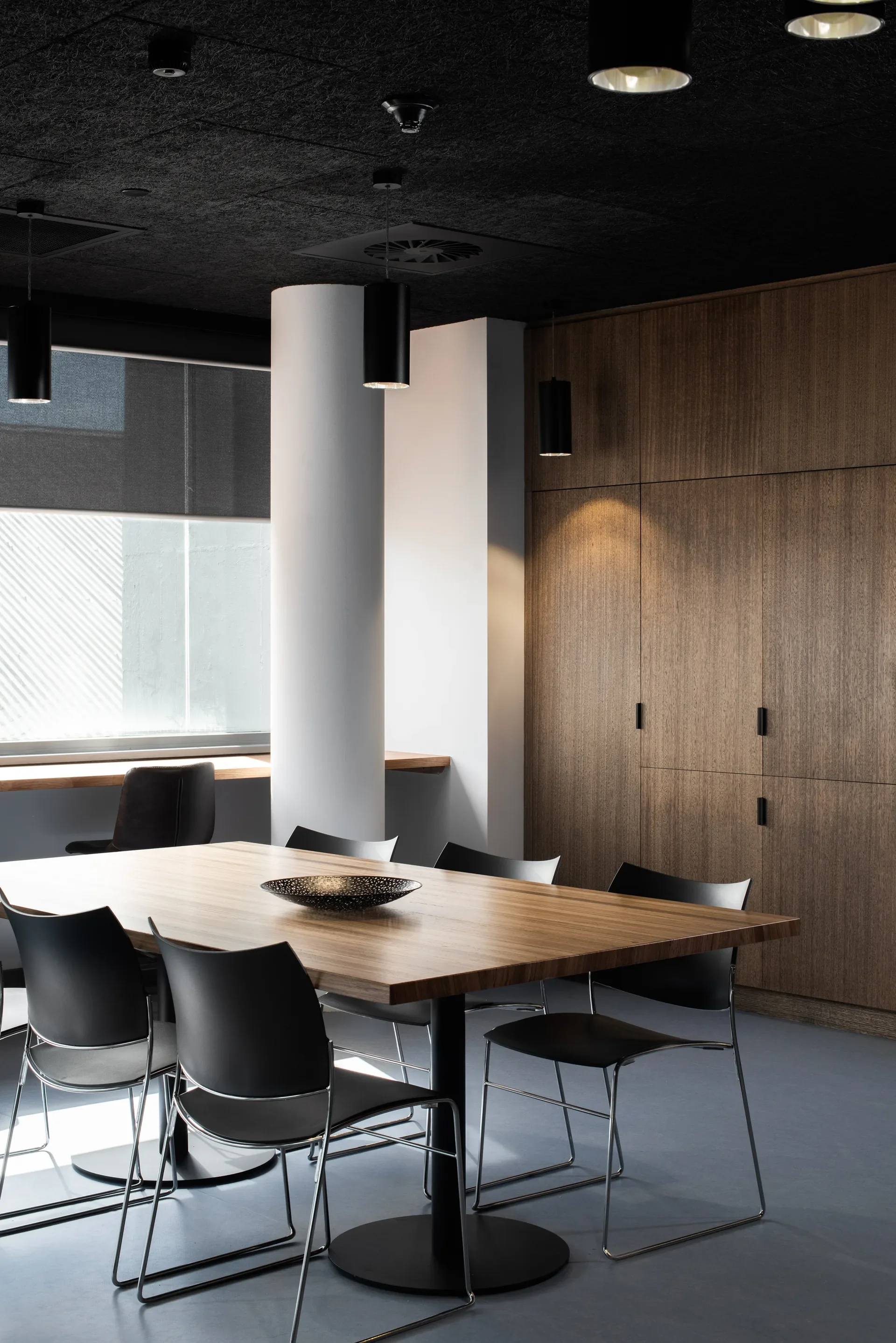 Black chairs surround a wooden table in a room featuring floor-to-ceiling timber cabinetry and a large white structural pillar. Cylindrical pendant lights hang from a dark, textured ceiling.
