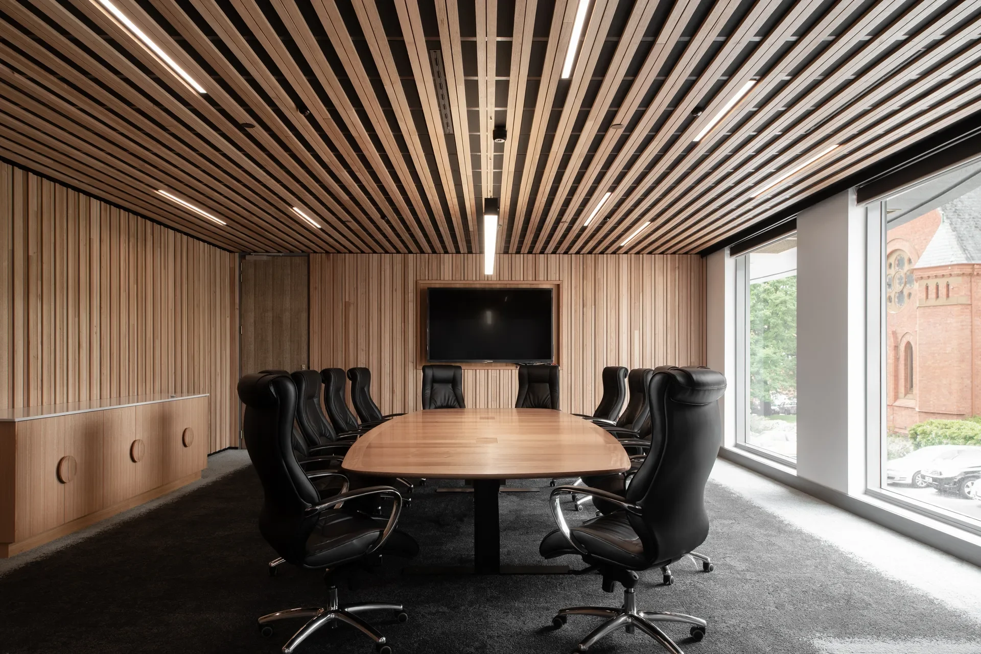 High-backed black chairs surround a wooden table in a boardroom featuring slatted timber walls and a matching ceiling with linear lights. A large window overlooks the same red-brick building seen from the lounge.