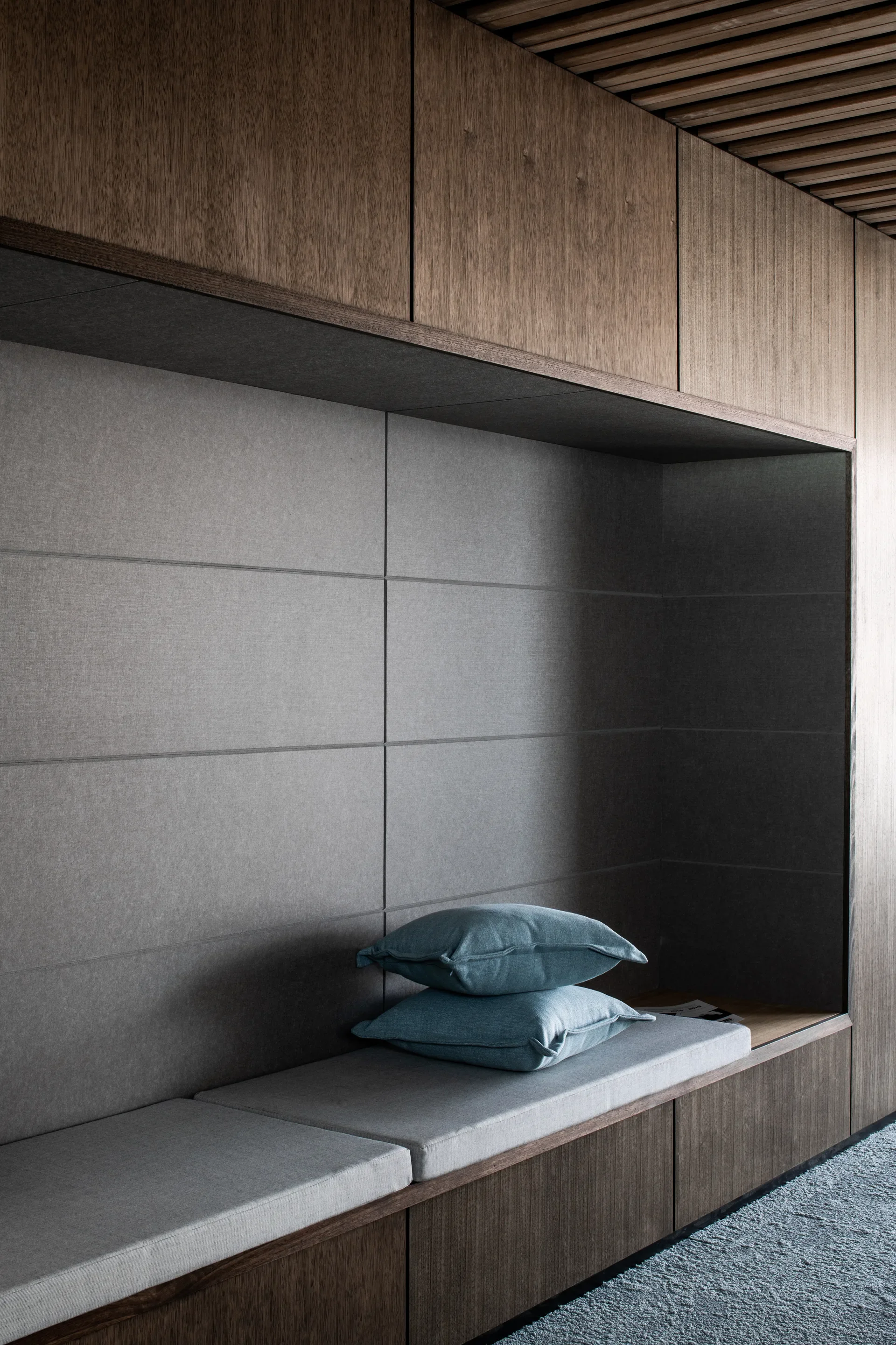 A cushioned bench is built into a timber-panelled alcove with grey fabric wall tiles and blue pillows. Overhead, the wooden panels meet a slatted timber ceiling.