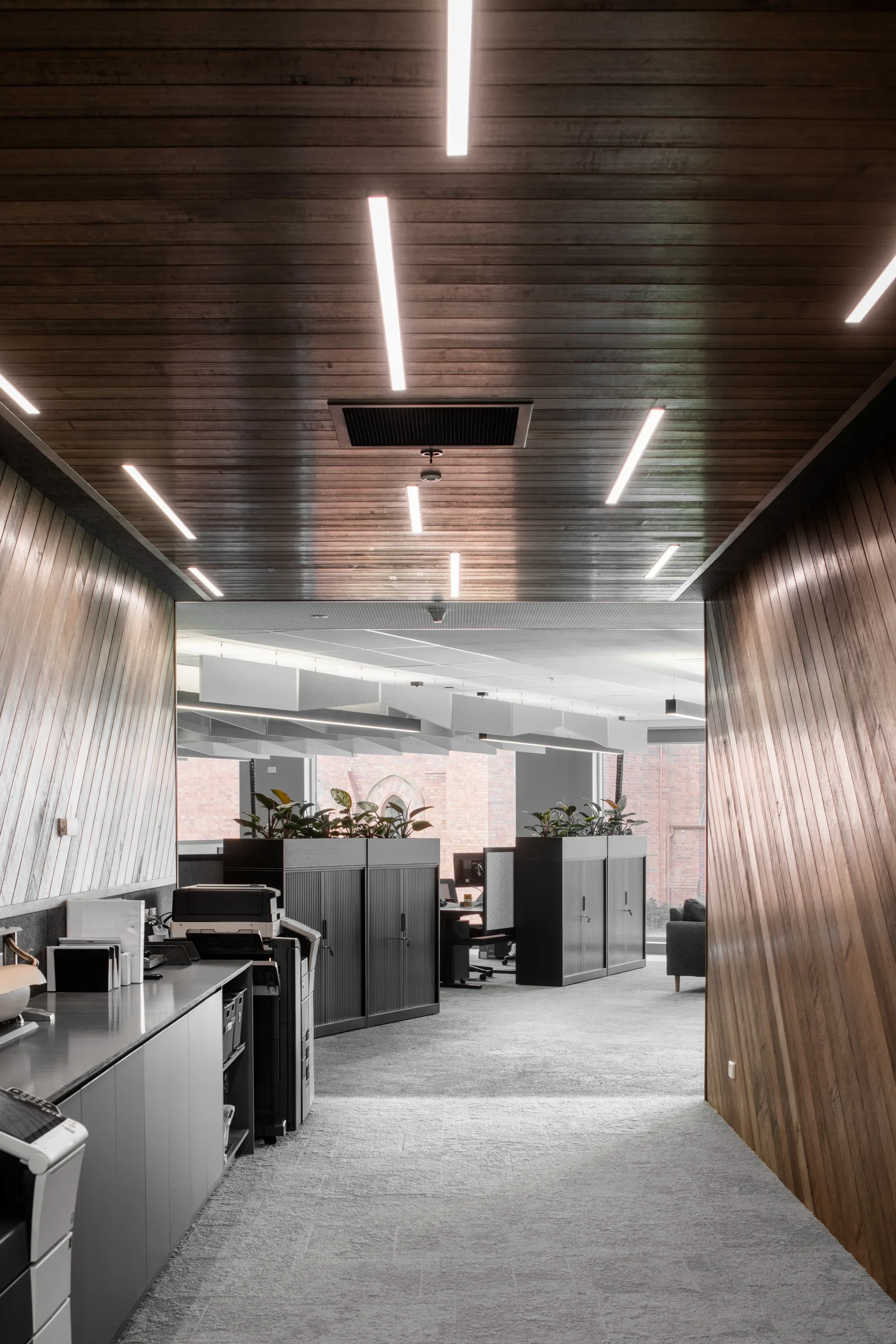 A hallway with diagonally slatted timber walls and matching recessed lighting leads past a printer station toward an open office. Black planter cabinets separate the corridor from the main workspace.