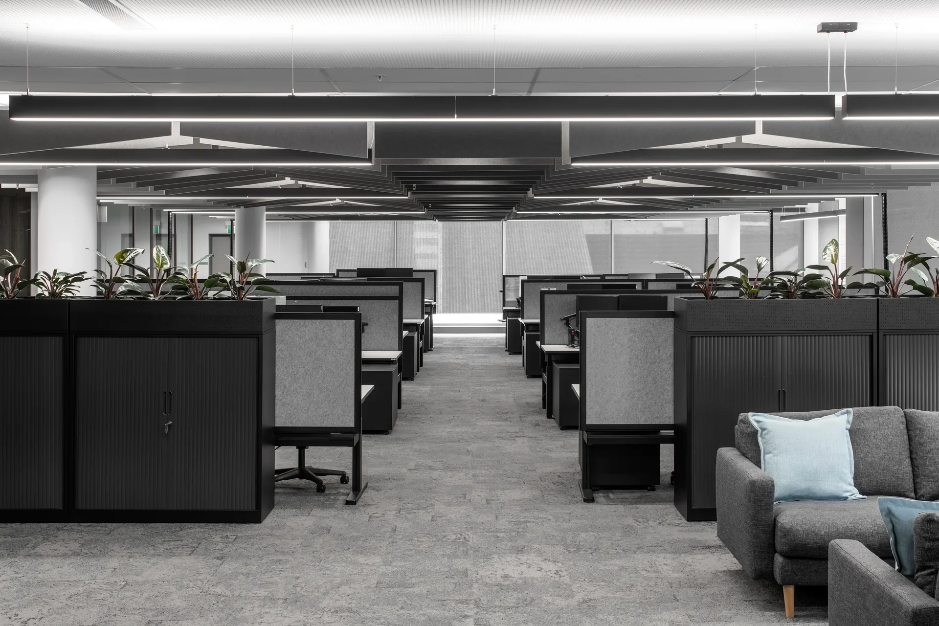 Rows of grey workstations are separated by black cabinets with integrated planters in a modern, open-plan office. Above, geometric acoustic baffles and linear pendant lights create a tiered pattern across the ceiling.