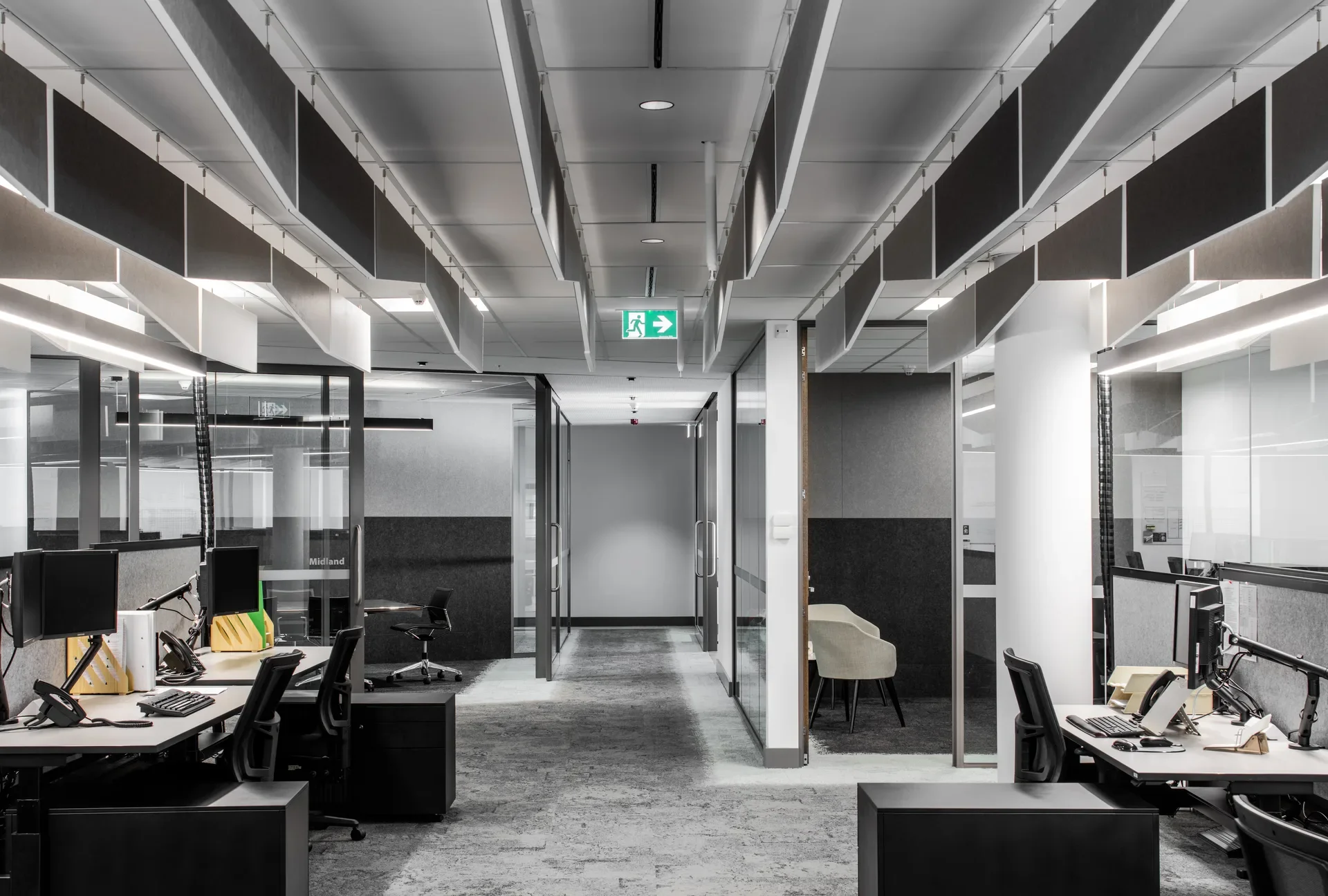 An alternate view of the office highlights a corridor with grey carpet that leads past glass-walled offices and workstations under a ceiling of linear lights and grey acoustic baffles.