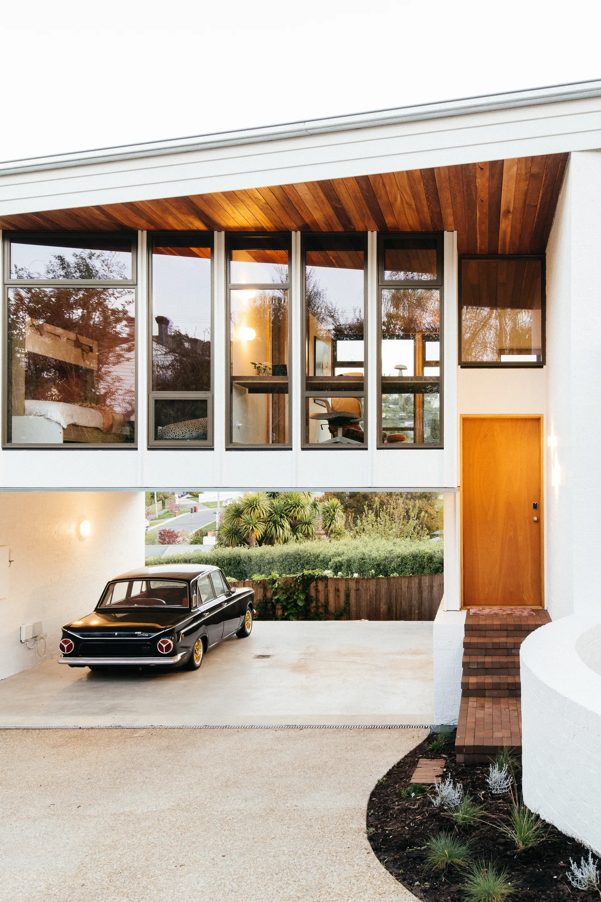 Mid-century modern home featuring a white exterior, timber ceilings, and a wood-slat door. A vintage black car is parked in a carport beneath a large, glass-walled upper level.