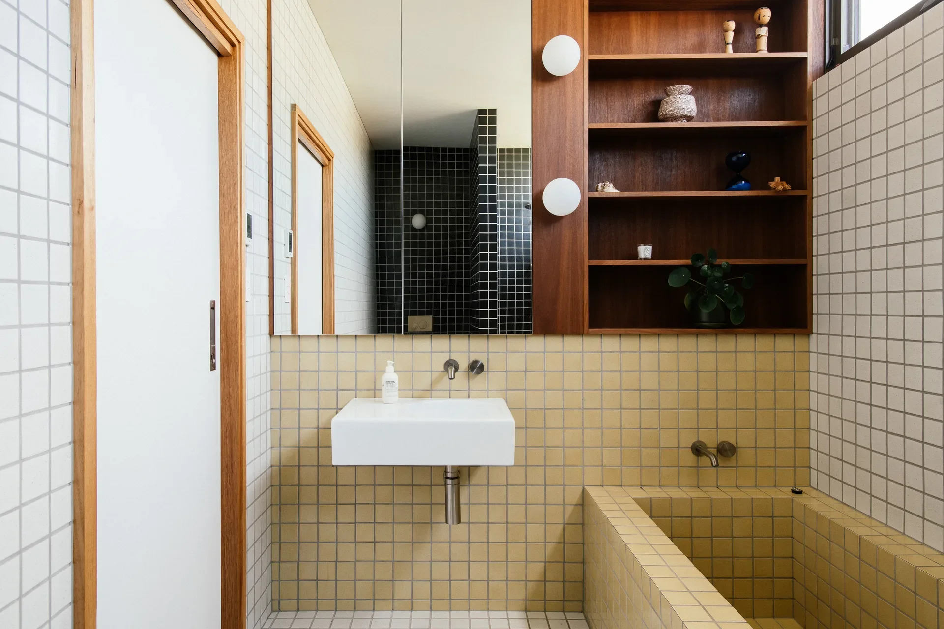 A bathroom featuring yellow and white grid tiling, a floating white sink, and a built-in tiled bathtub. A dark wood shelving unit and large mirror add warmth and depth to the minimalist design.
