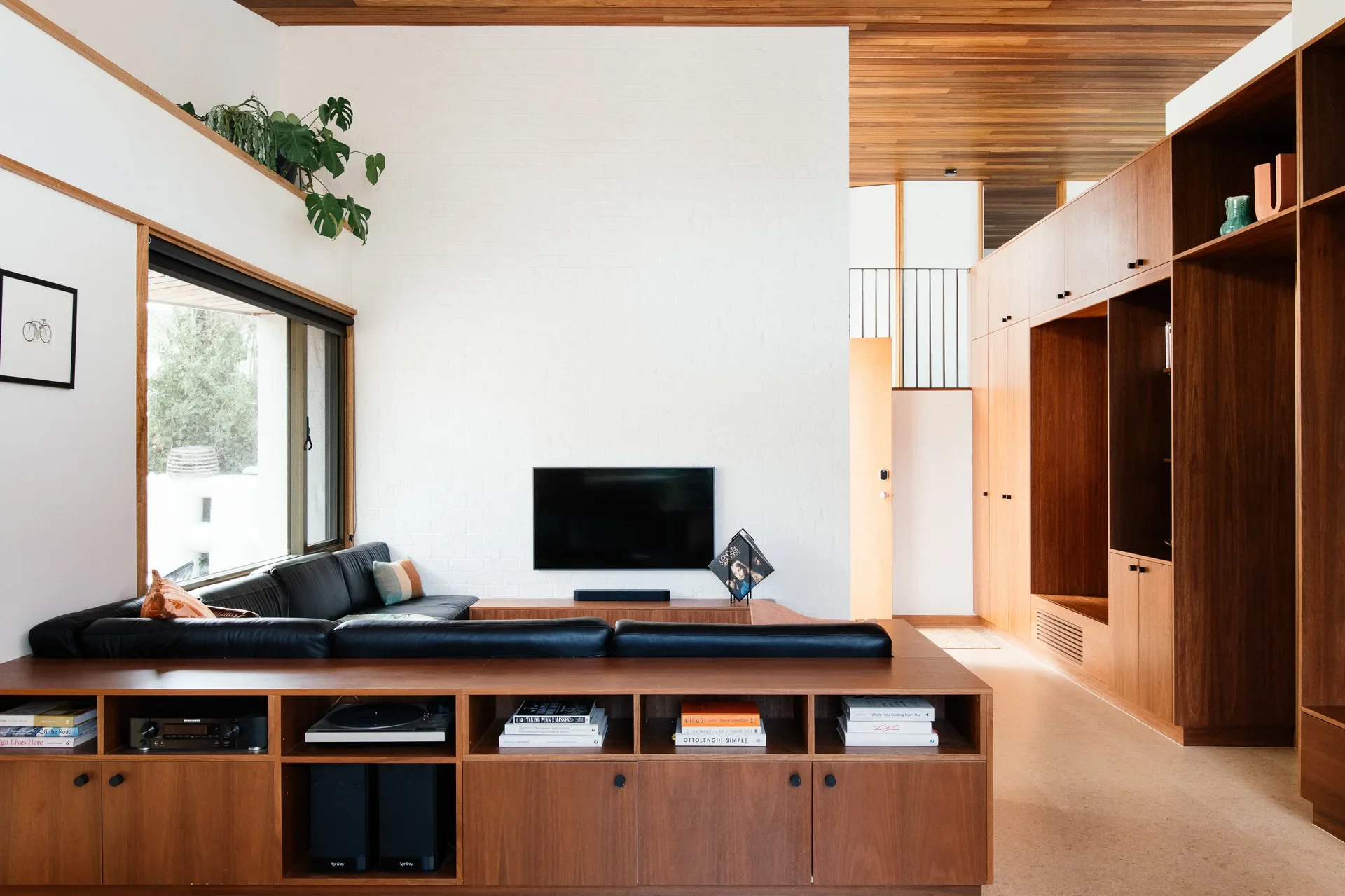 A living room featuring a black leather sofa integrated with custom wood shelving. The space is defined by bright white walls, a slatted timber ceiling, and expansive warm wood cabinetry that offers plenty of storage and display space.