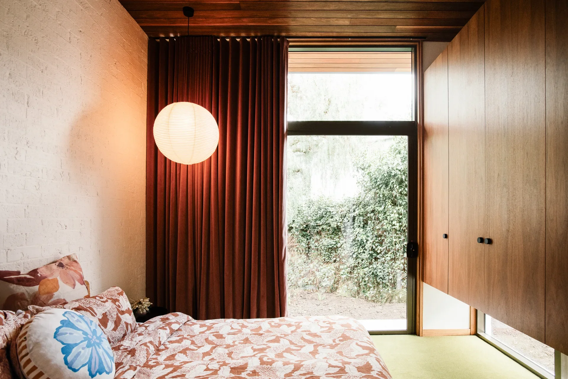 Cozy bedroom featuring patterned floral bedding, a hanging paper globe lamp, and deep burgundy curtains. A large glass door opens to a garden, while floating wood cabinets and a timber ceiling add a warm, mid-century feel.