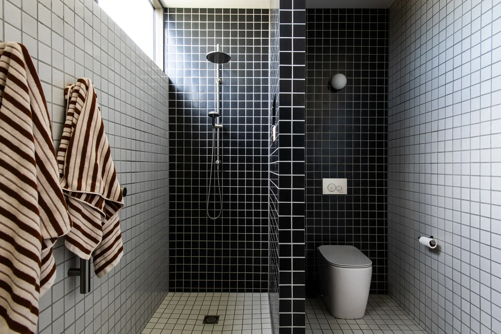 A bathroom with black and grey grid tiling, featuring a walk-in shower and a minimalist toilet area. Striped towels hang on the left, adding texture to the clean, geometric design.