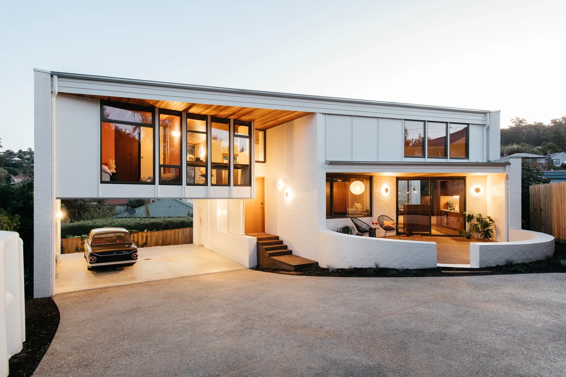 An expansive view of the mid-century modern white house at dusk. Warm interior lighting glows through the large glass windows of the cantilevered upper level and the curved patio below.