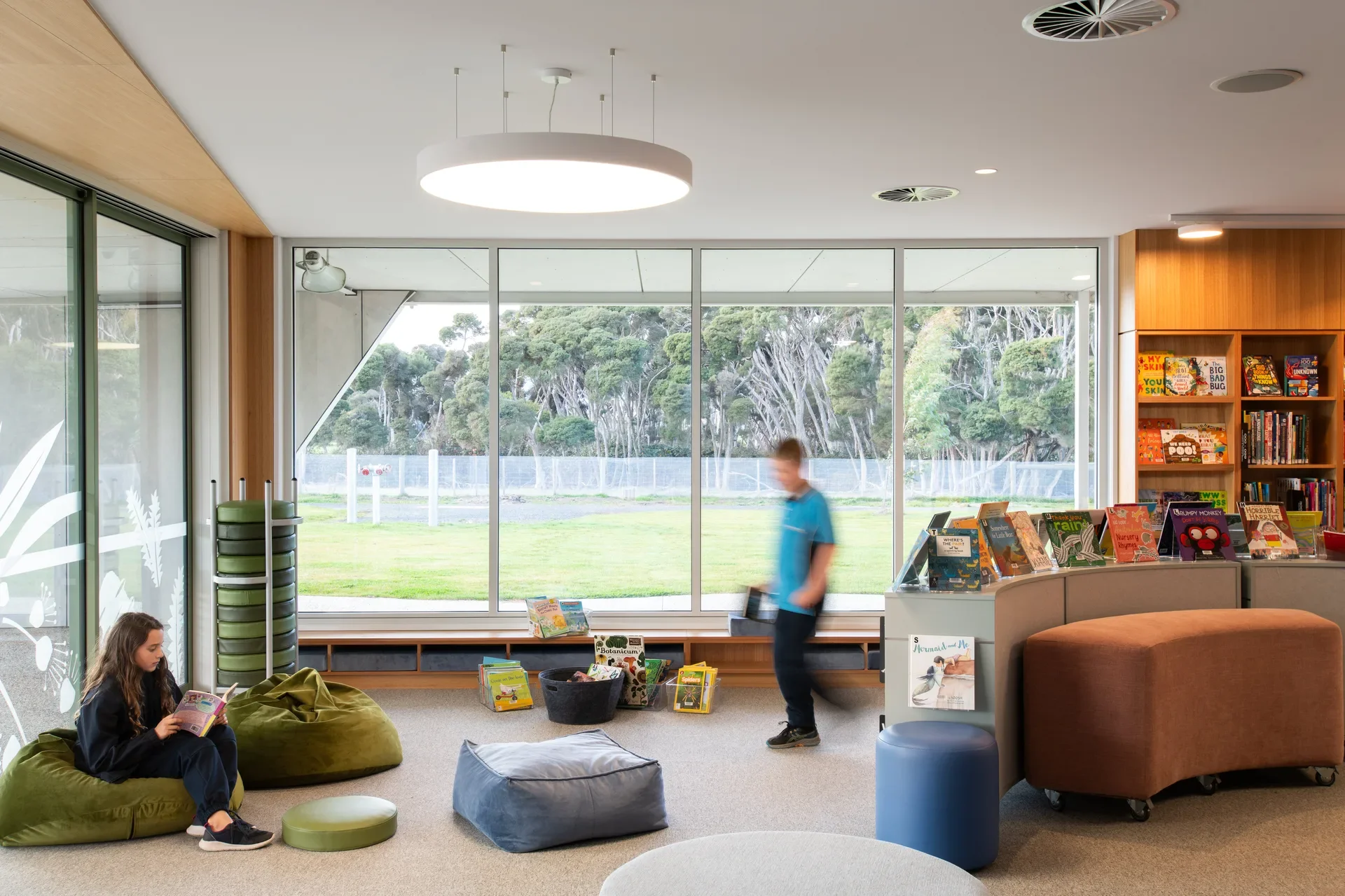 This bright reading area features large floor-to-ceiling windows overlooking a grassy field and a line of trees. Inside, children are using green and blue beanbag chairs and curved benches near low bookshelves filled with colourful books.
