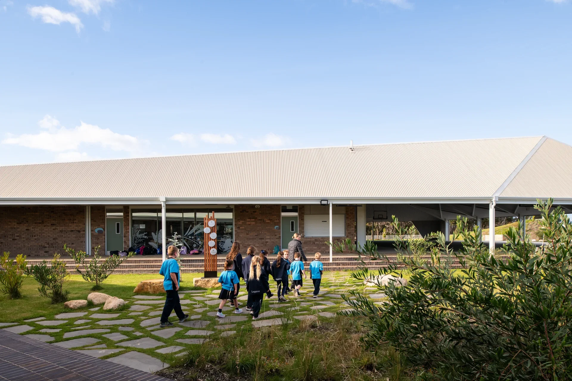 Gemini said This building features a long, sloped corrugated roof and brick exterior walls. A group of children walk along a stone-paved path through a grassy lawn toward the building.
