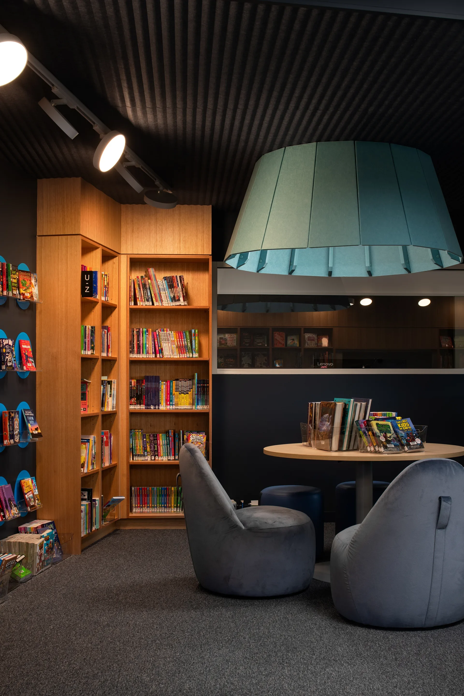 This cozy reading nook features wooden bookshelves and a small round table surrounded by grey beanbag chairs. A large, tapered teal pendant light hangs from a dark, corrugated ceiling, providing focused illumination over the space.