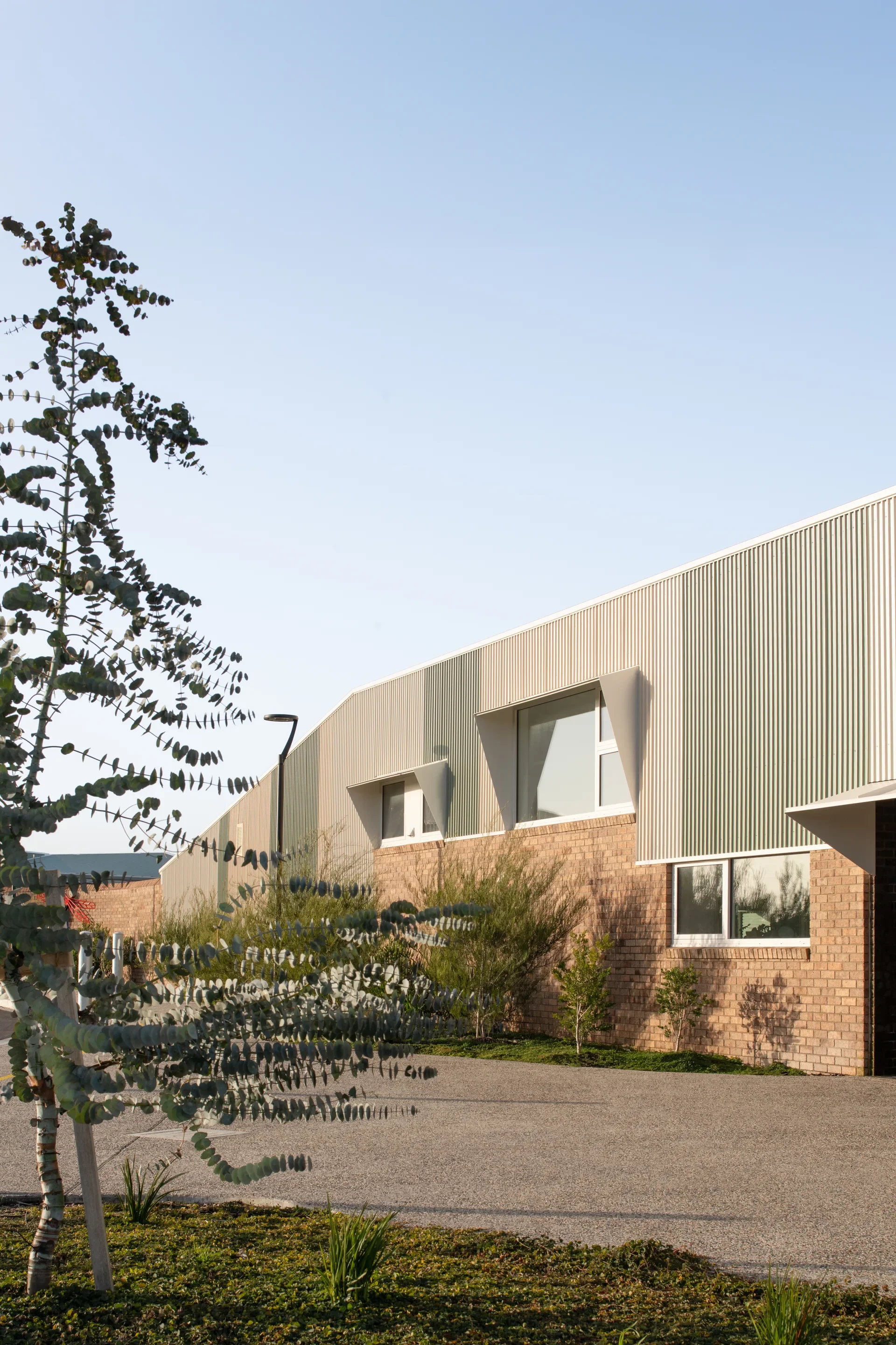 Gemini said This building features a mix of tan brick and vertical corrugated metal siding in varying shades of green and grey. Several windows are set into the upper section, some highlighted by angled metallic hoods that provide shade and architectural interest.