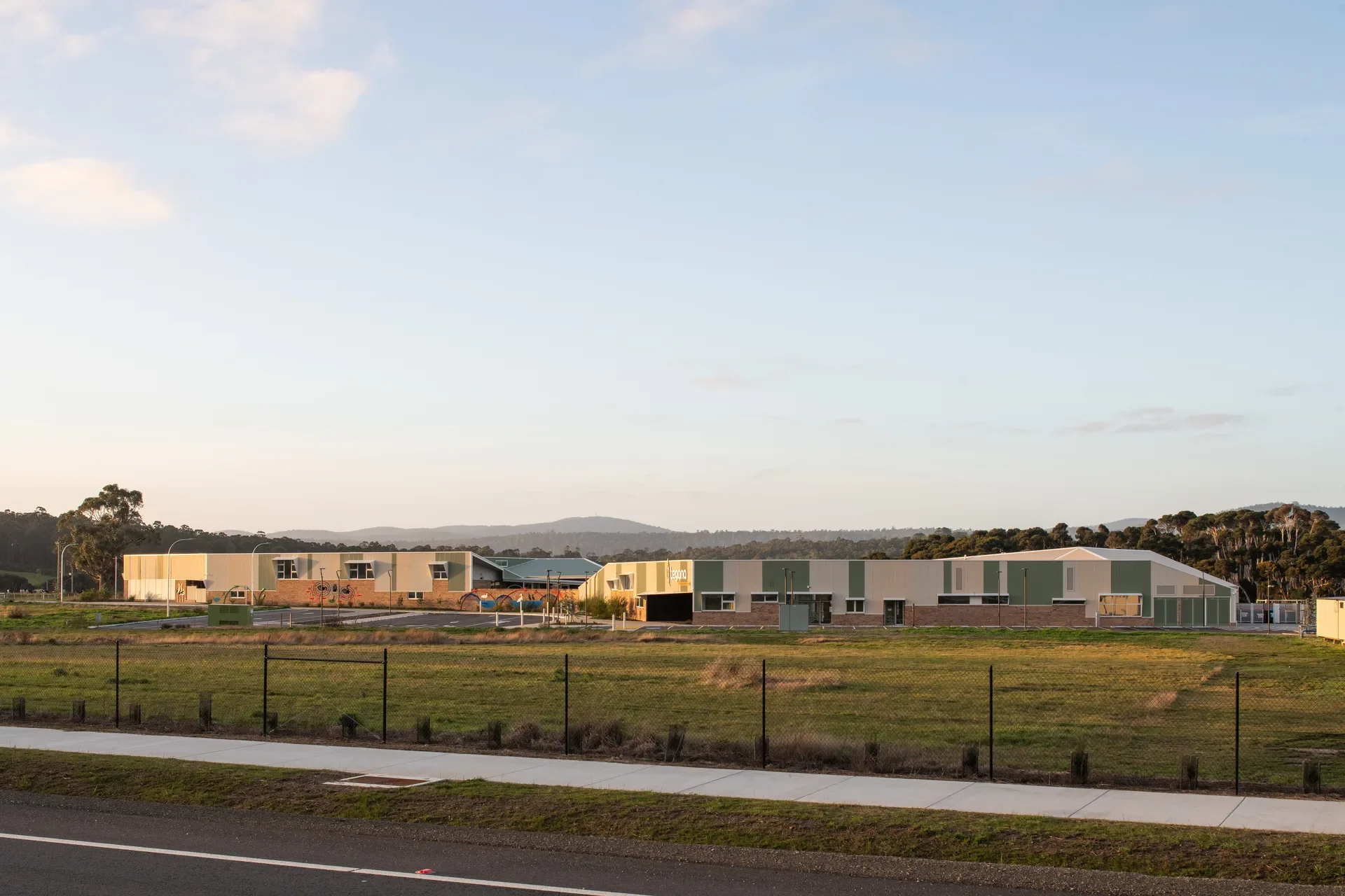 This wide-angle view shows the modern school facility situated in a rural landscape with distant hills. The buildings feature green and tan corrugated siding and low-profile roofs, bordered by a large grassy field and a paved road in the foreground.