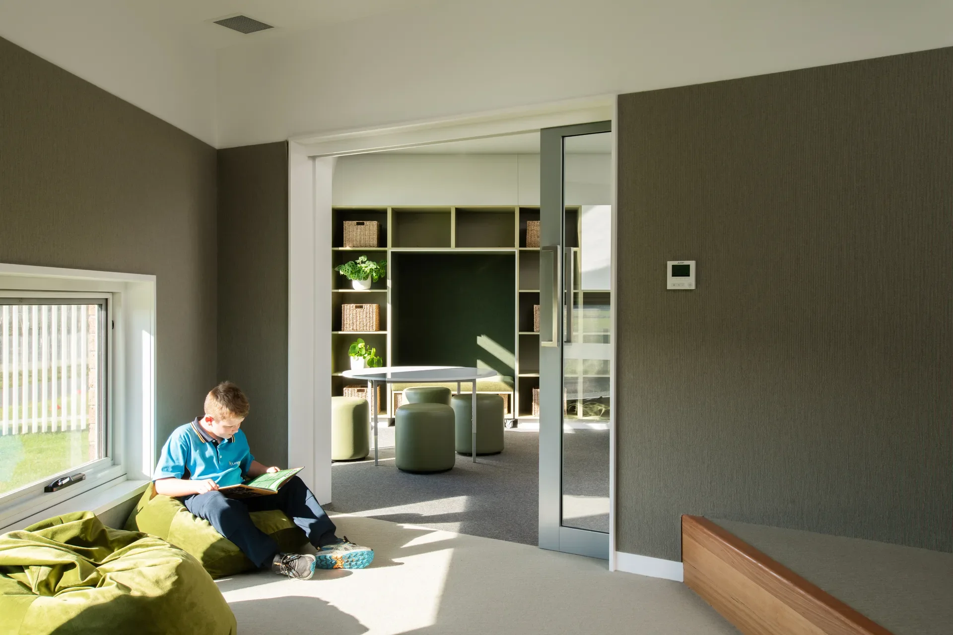 Gemini said In this indoor setting, a child sits on a large green beanbag while reading a book. A sliding glass door opens into an adjacent room featuring olive green walls, matching cylindrical stools, and shelving units filled with woven baskets.