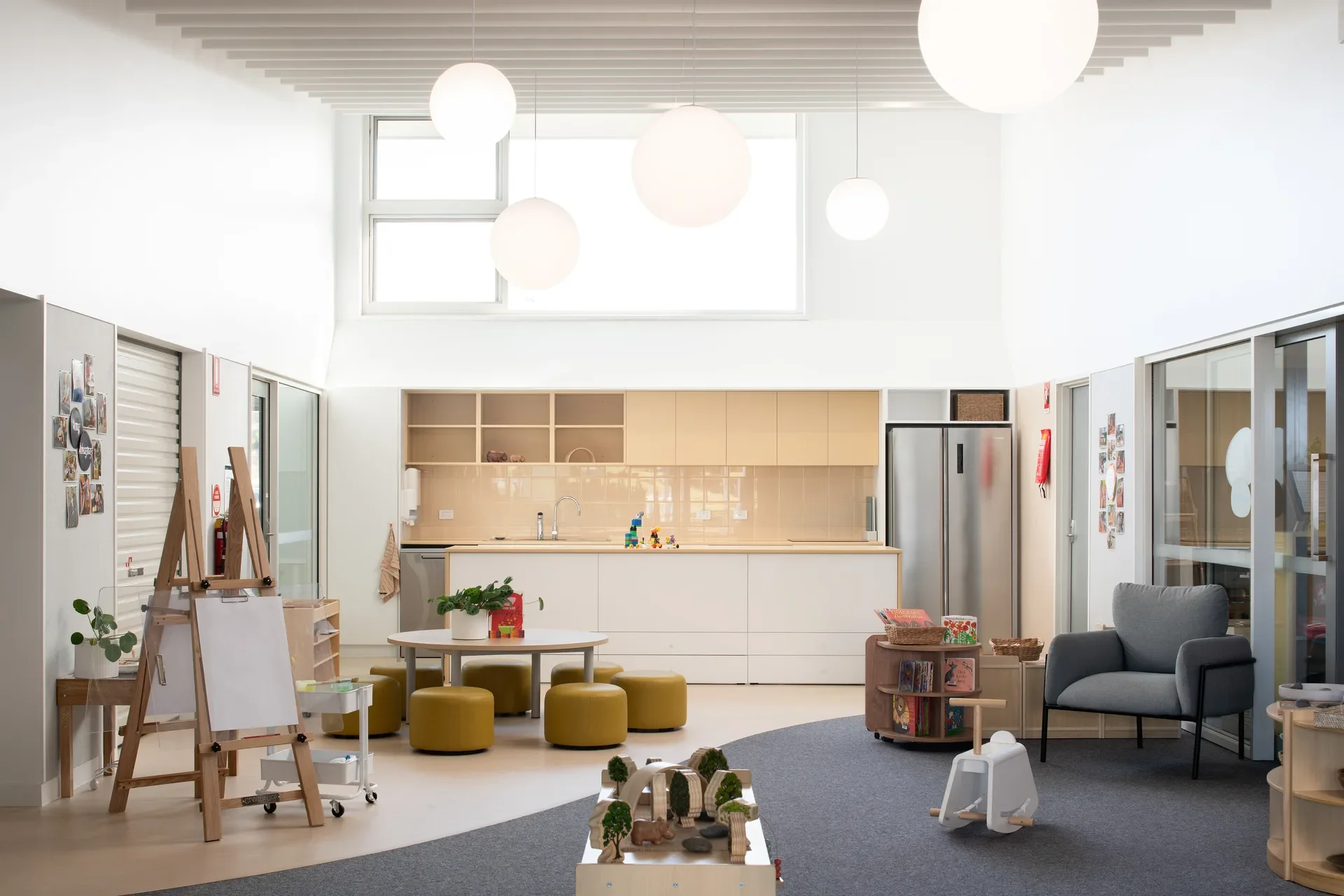 This bright communal space features a modern kitchen with light wood cabinetry and a white island. Large spherical pendant lights hang above a play area equipped with an art easel, a toy train set, and small yellow cylindrical stools.