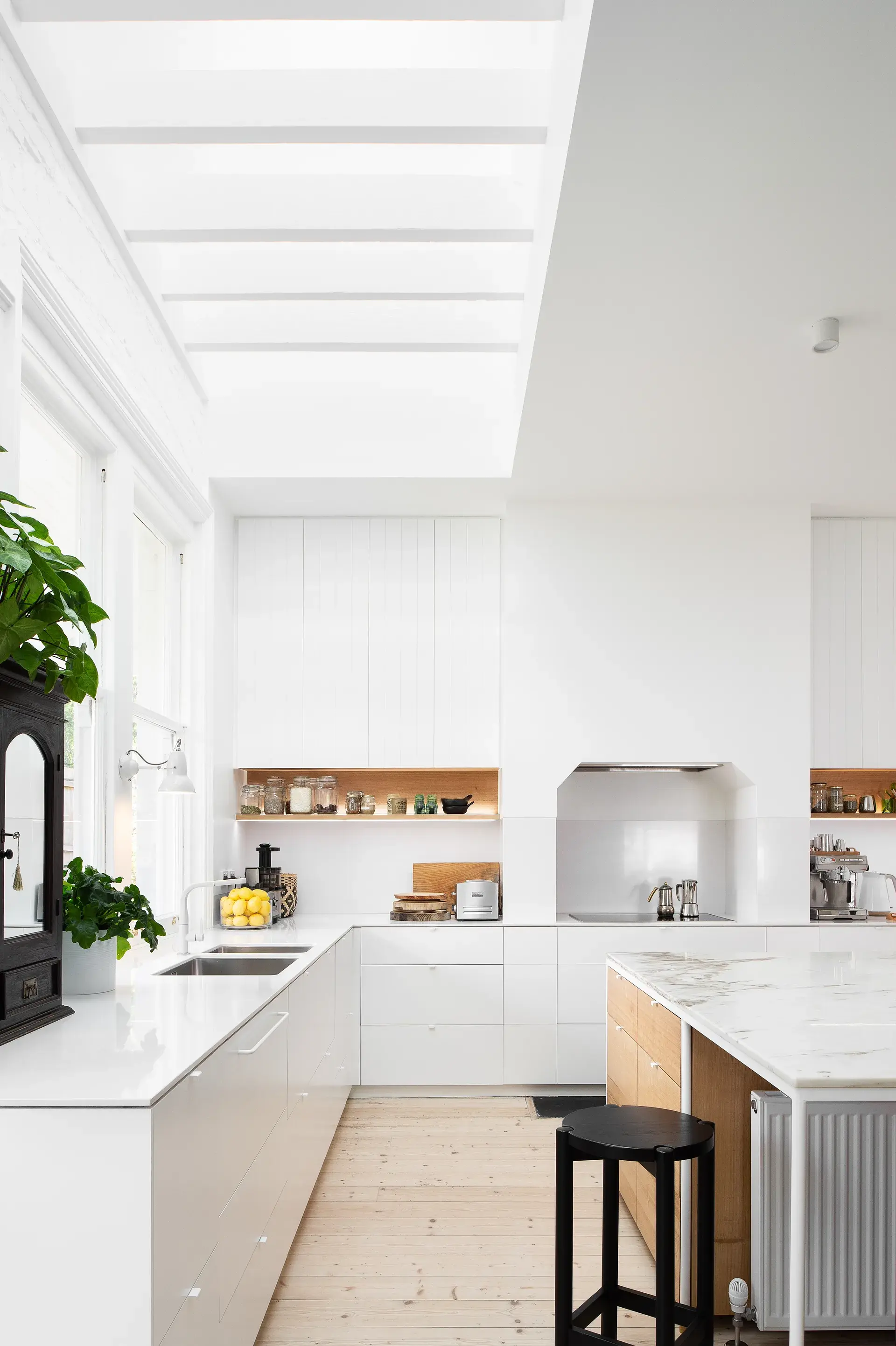 This ultra-modern kitchen features an all-white design with a large marble-topped island and light wood flooring. A high, slatted skylight floods the space with natural light, highlighting the minimalist white cabinetry and integrated appliances.