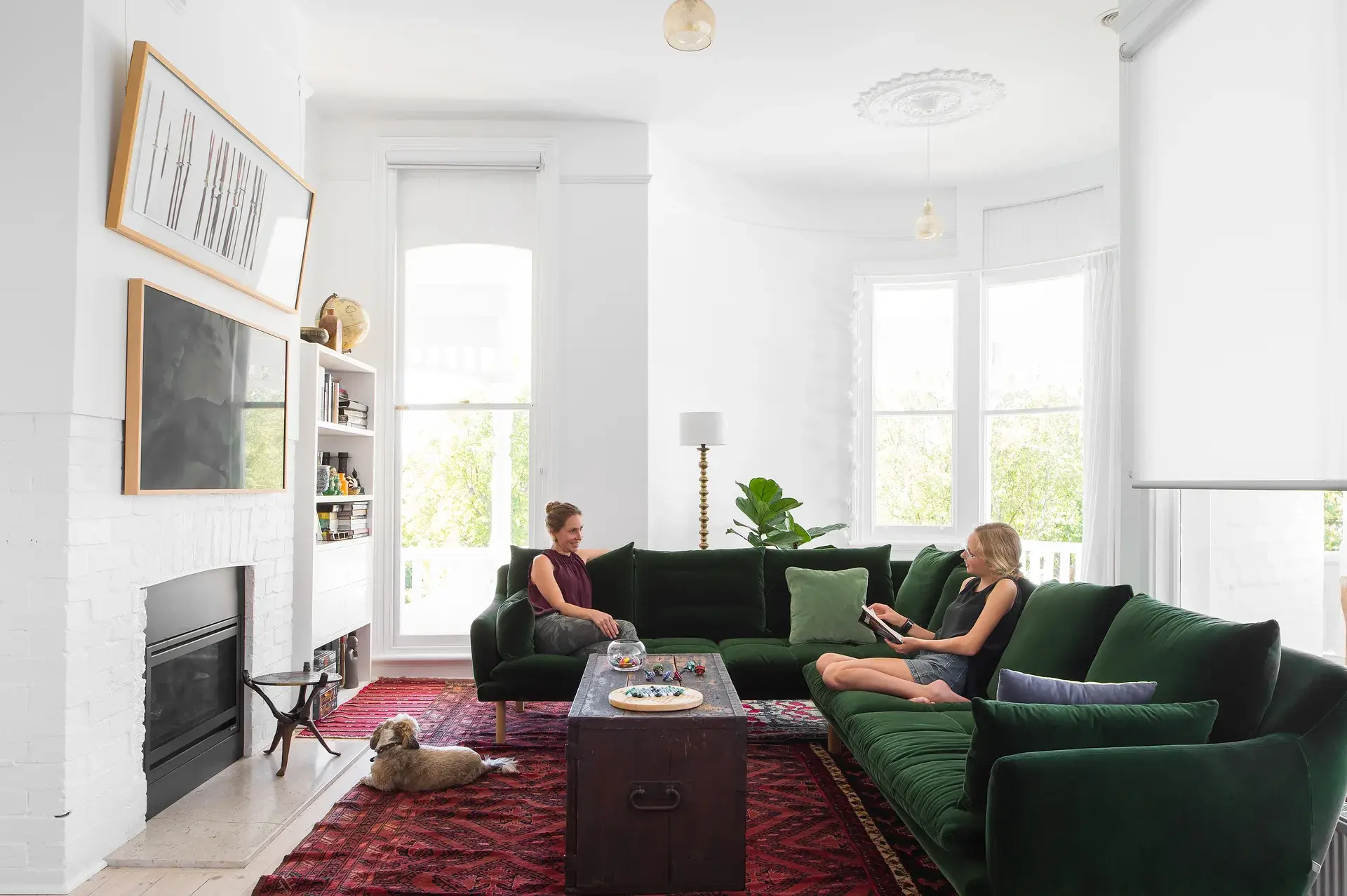 This bright living room features a large, green velvet sofa and a dark wooden trunk used as a coffee table. Two people relax on the sofa over a red patterned rug, while a small dog rests on the floor near a white brick fireplace.