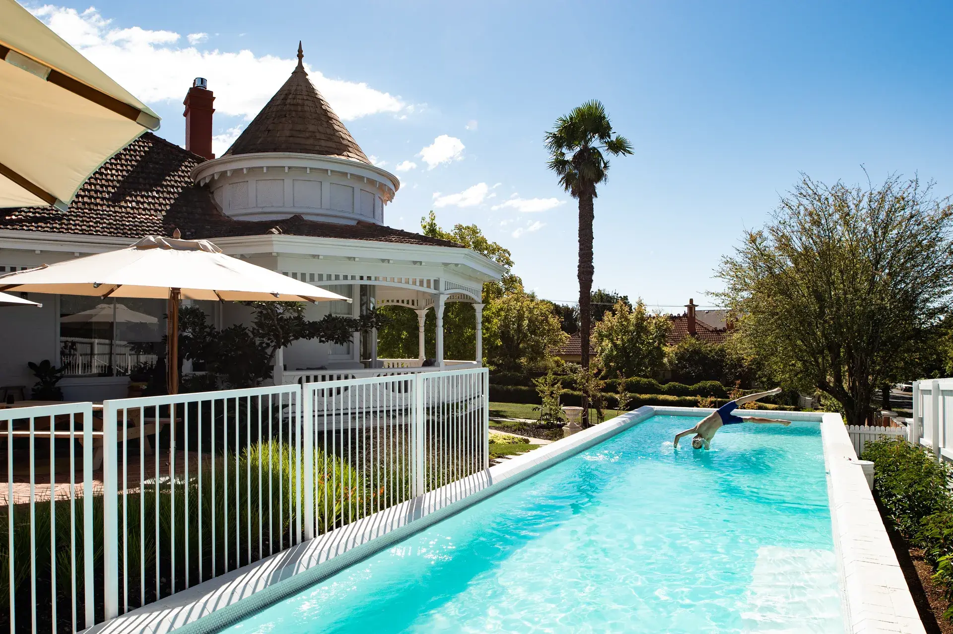 This outdoor scene features a long, narrow swimming pool with a white safety fence. A person is mid-dive into the clear blue water, while a traditional white house with a wooden turret and a tall palm tree stand in the background under a bright, sunny sky.