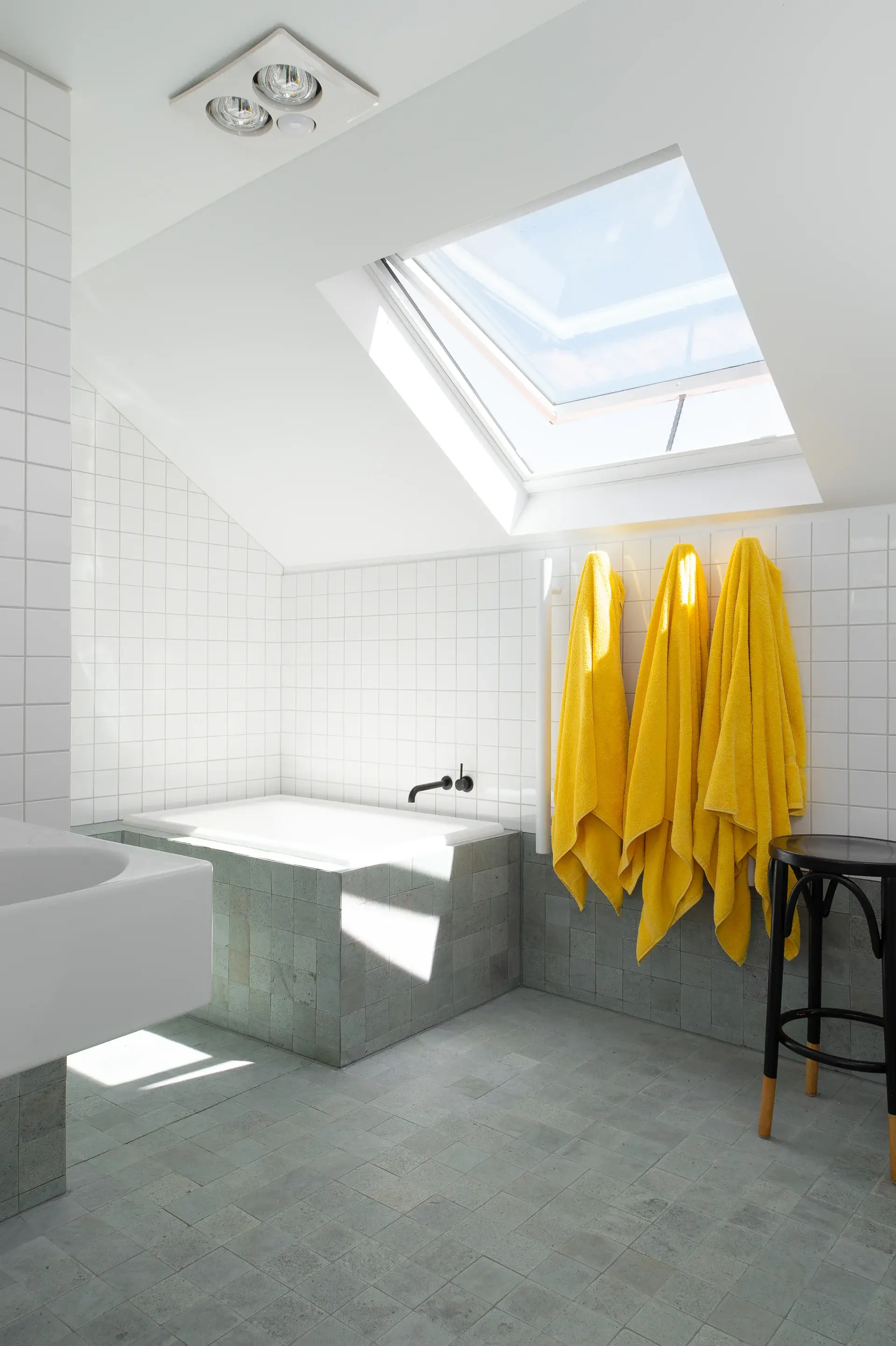 This modern bathroom features a sunken bathtub and grey stone floor tiles. Three bright yellow towels hang on a white tiled wall beneath a large skylight that floods the room with natural light.