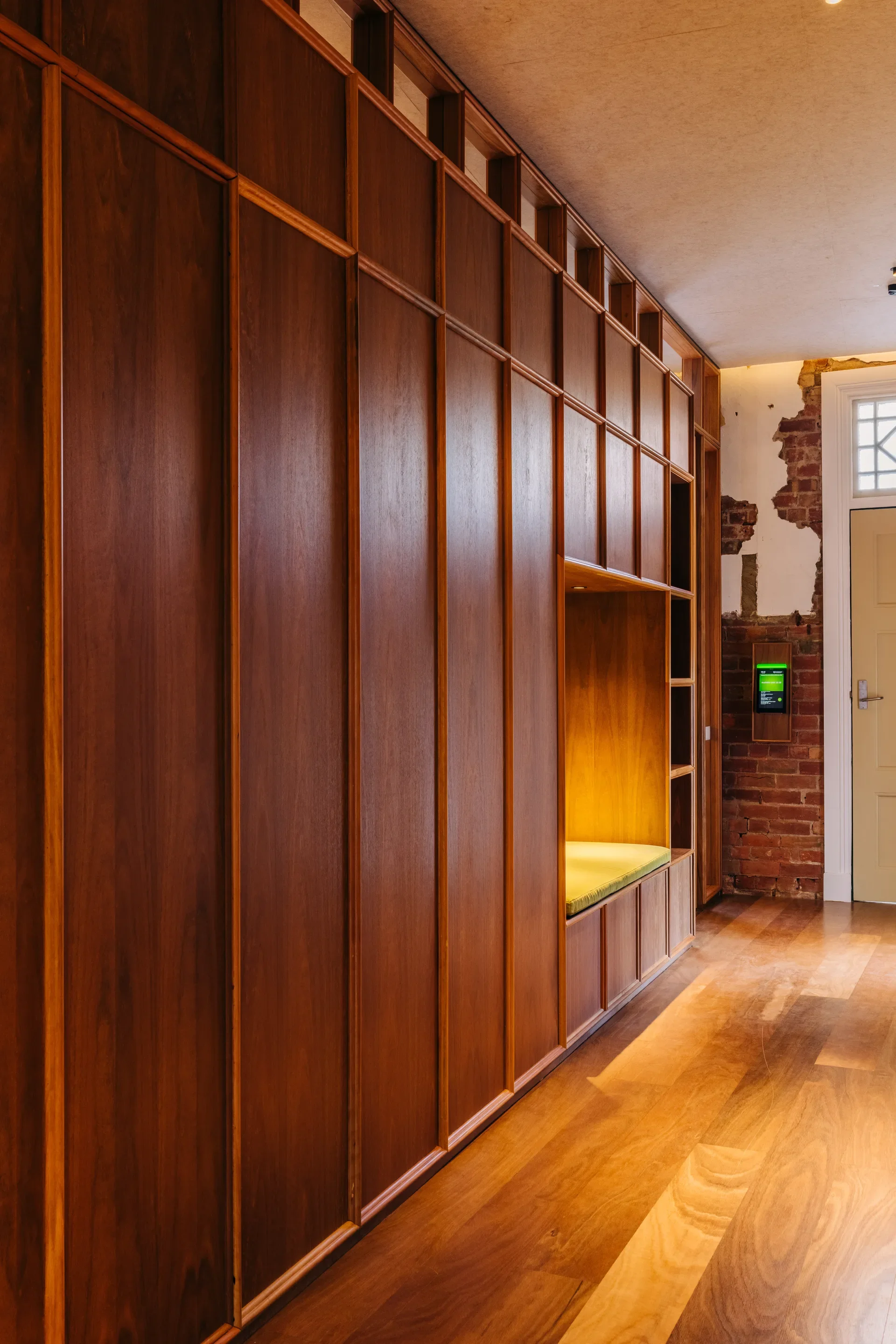 A hallway featuring a floor-to-ceiling wooden storage wall with a built-in seating nook. The warm wood tones contrast with an exposed red brick wall at the end of the corridor, creating a modern, textured look.