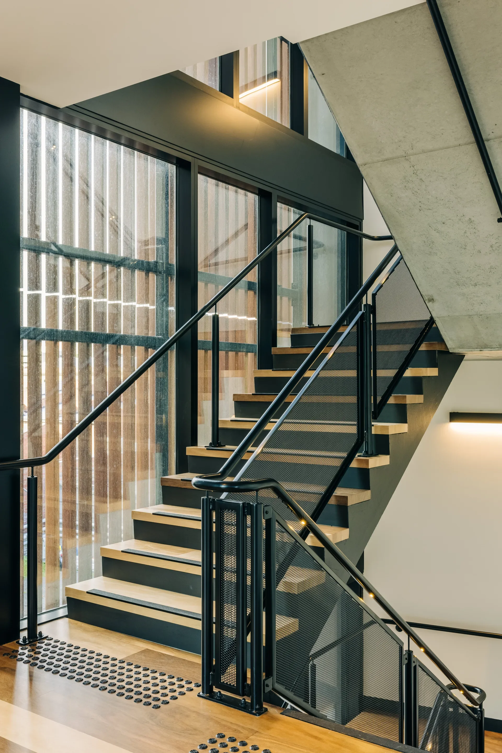 Modern staircase featuring timber treads and black mesh railings. The design blends industrial and contemporary elements with exposed concrete ceilings and large glass windows that filter in soft, natural light.