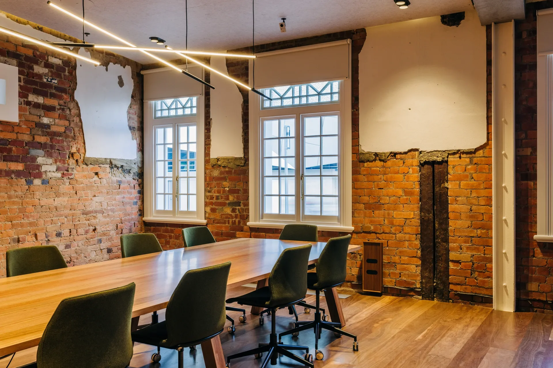 A conference room blending historic and modern styles, featuring a long timber table and green chairs. The space is defined by exposed red brick walls with patches of white plaster, classic white-framed windows, and striking linear pendant lighting.