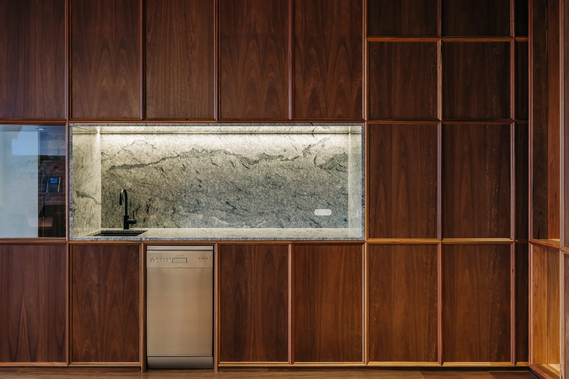 Minimalist kitchenette integrated into a rich wood-paneled wall. It features a marble backsplash with built-in lighting, a black faucet, and a sleek stainless steel dishwasher tucked neatly among the custom cabinetry.