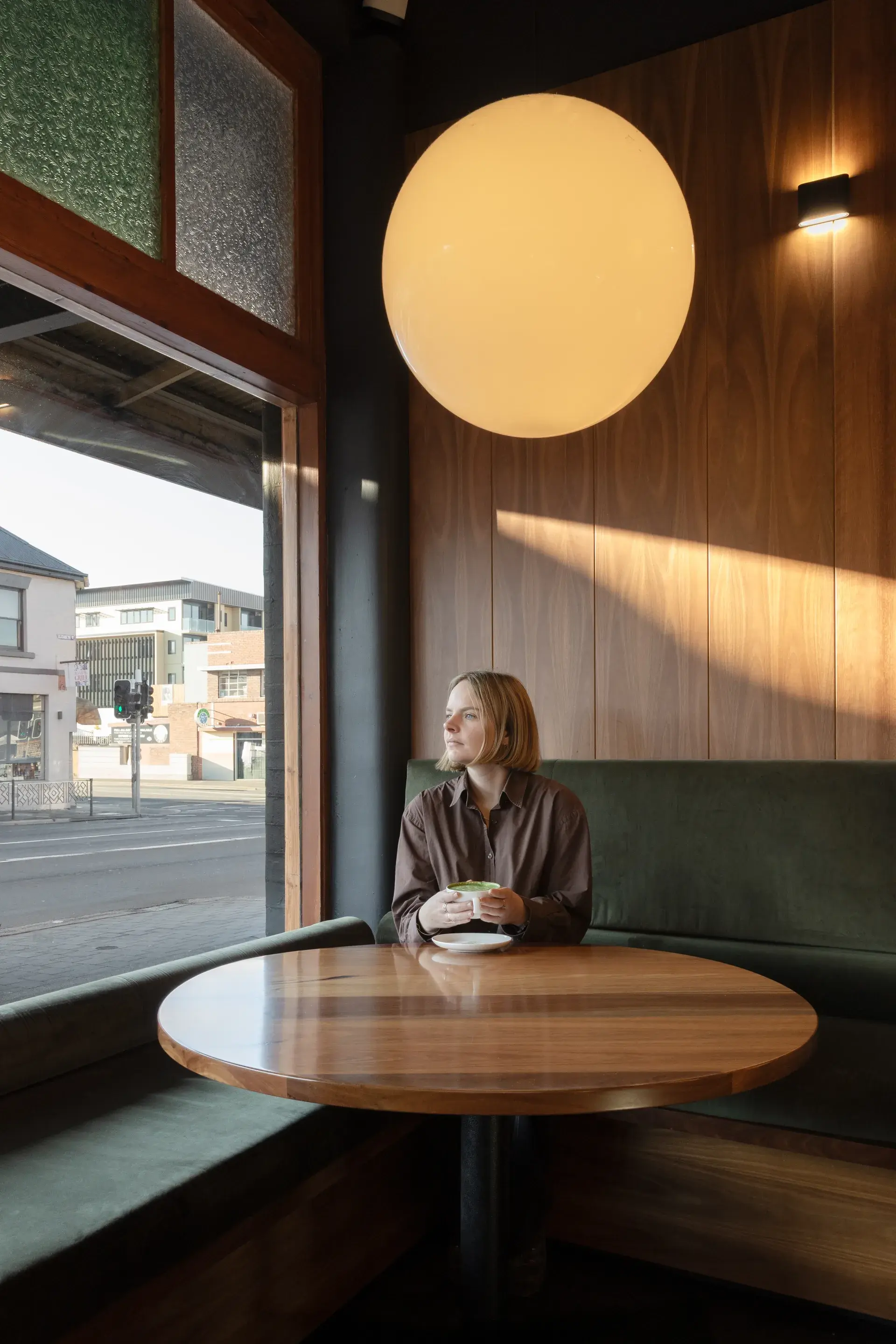 A large round timber table with green velvet banquette at the front of the restaurant, with a glass sphere pendant casting a warm glow above.