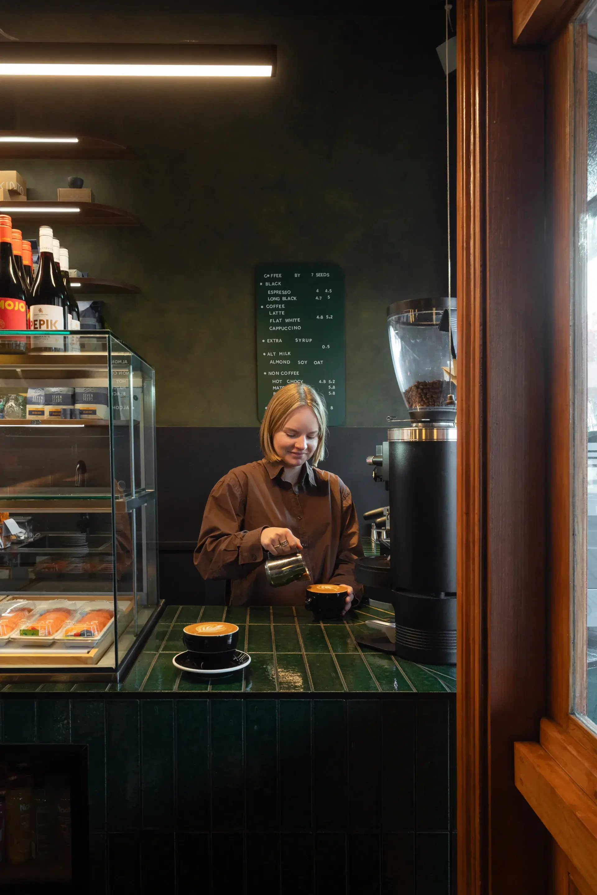 A barista prepares coffee behind the green tiled counter, with a rich Porter’s Paints French Wash feature wall in Seed and Antiquity beyond.