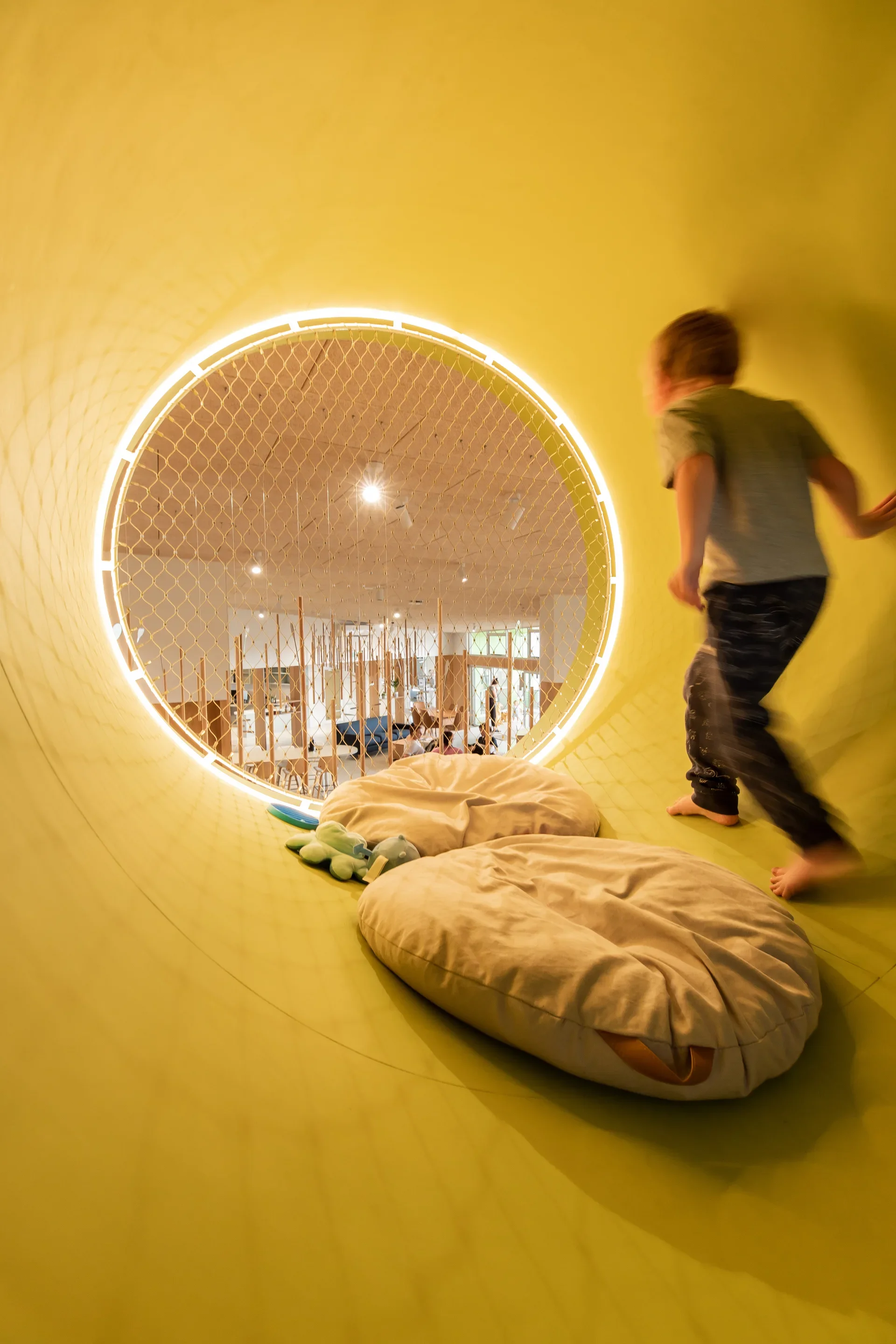 This interior view is taken from inside a bright yellow circular nook looking out into a larger play area. A child is seen moving within the yellow tunnel, which is furnished with soft beige beanbags and features a mesh-covered opening illuminated by a ring of light.