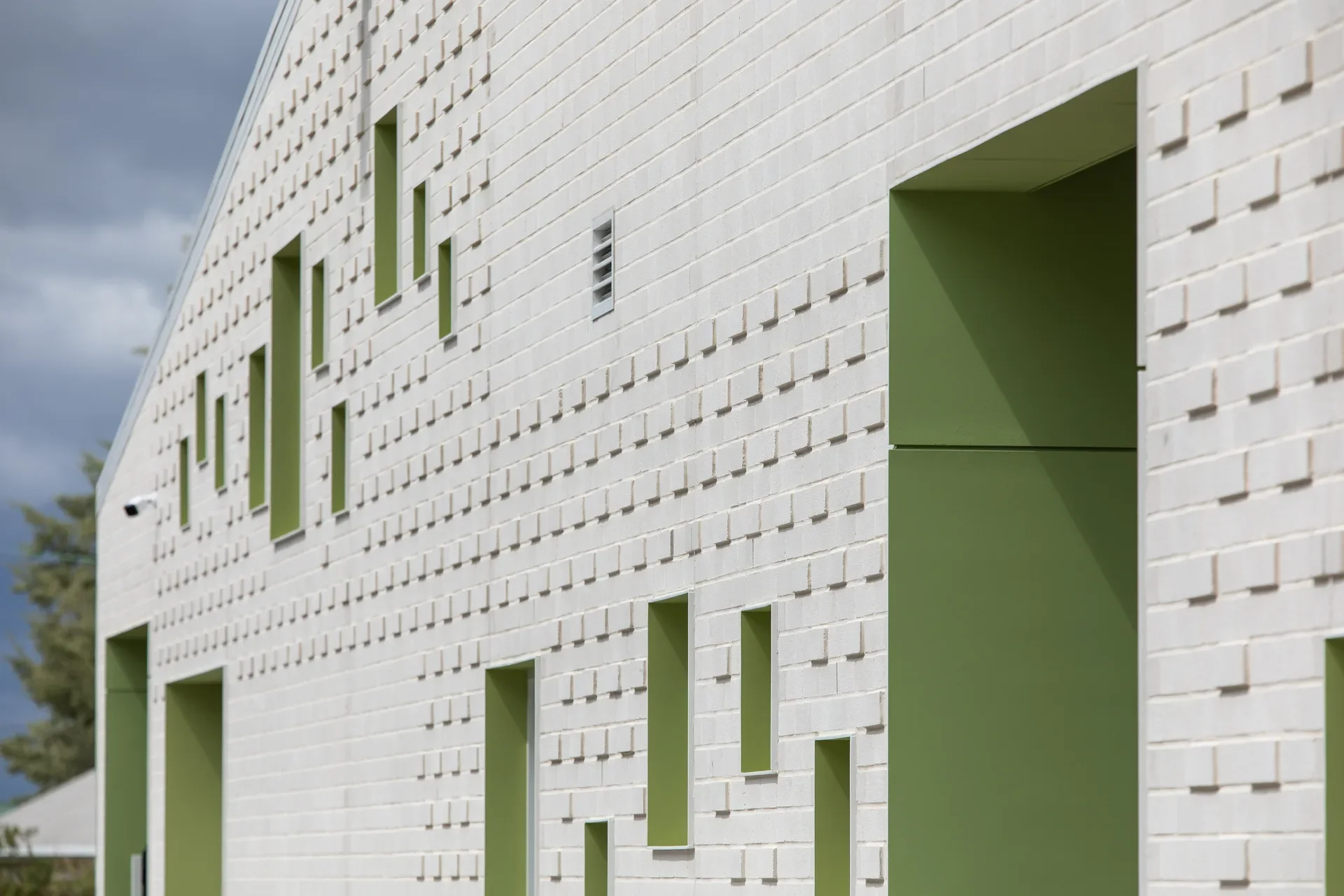 This white brick wall features a series of narrow, green-framed windows of varying heights. The facade is textured with a repetitive pattern of protruding bricks, and a large recessed entryway with matching green walls is visible in the foreground.