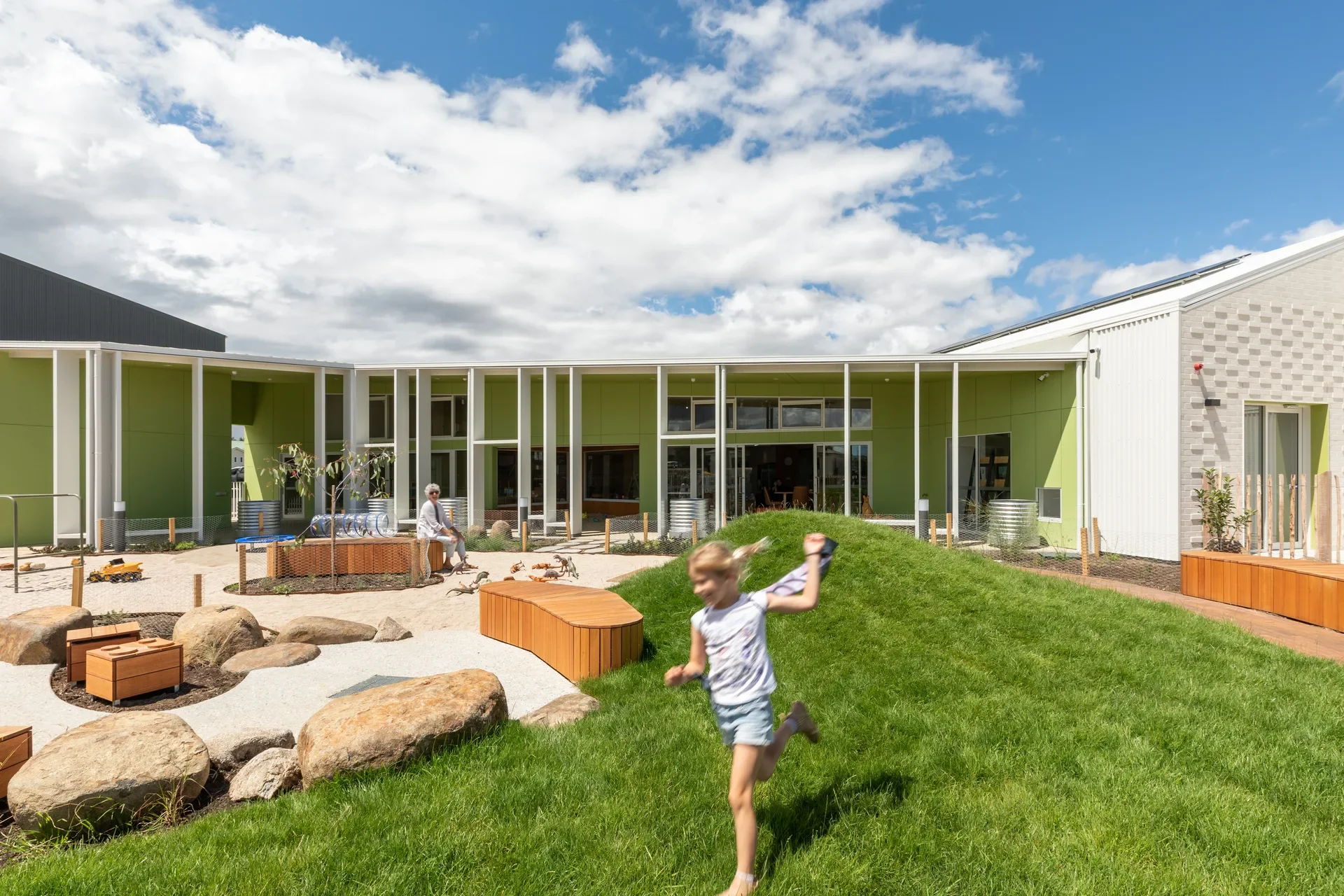 This outdoor play area features a grassy hill where a child is running, situated next to a sandpit with large rocks and wooden play structures. The background shows a modern building with vibrant lime green walls and a covered walkway supported by white pillars under a bright, partly cloudy sky.