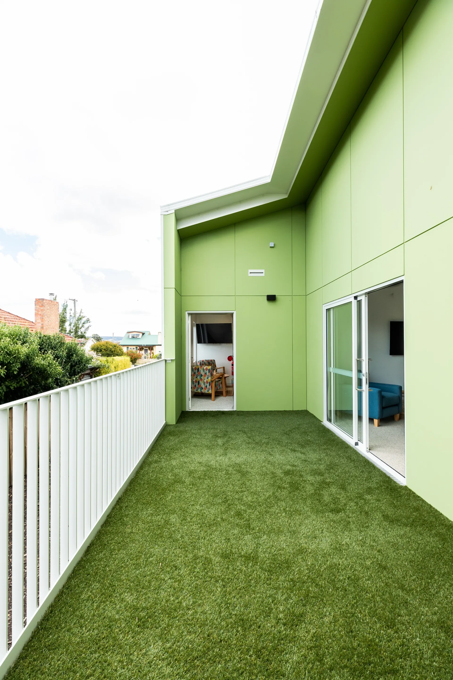 This narrow outdoor balcony features vibrant lime green walls and a floor covered in artificial turf. A white vertical railing runs along the left side, and a sliding glass door leads into an interior room.