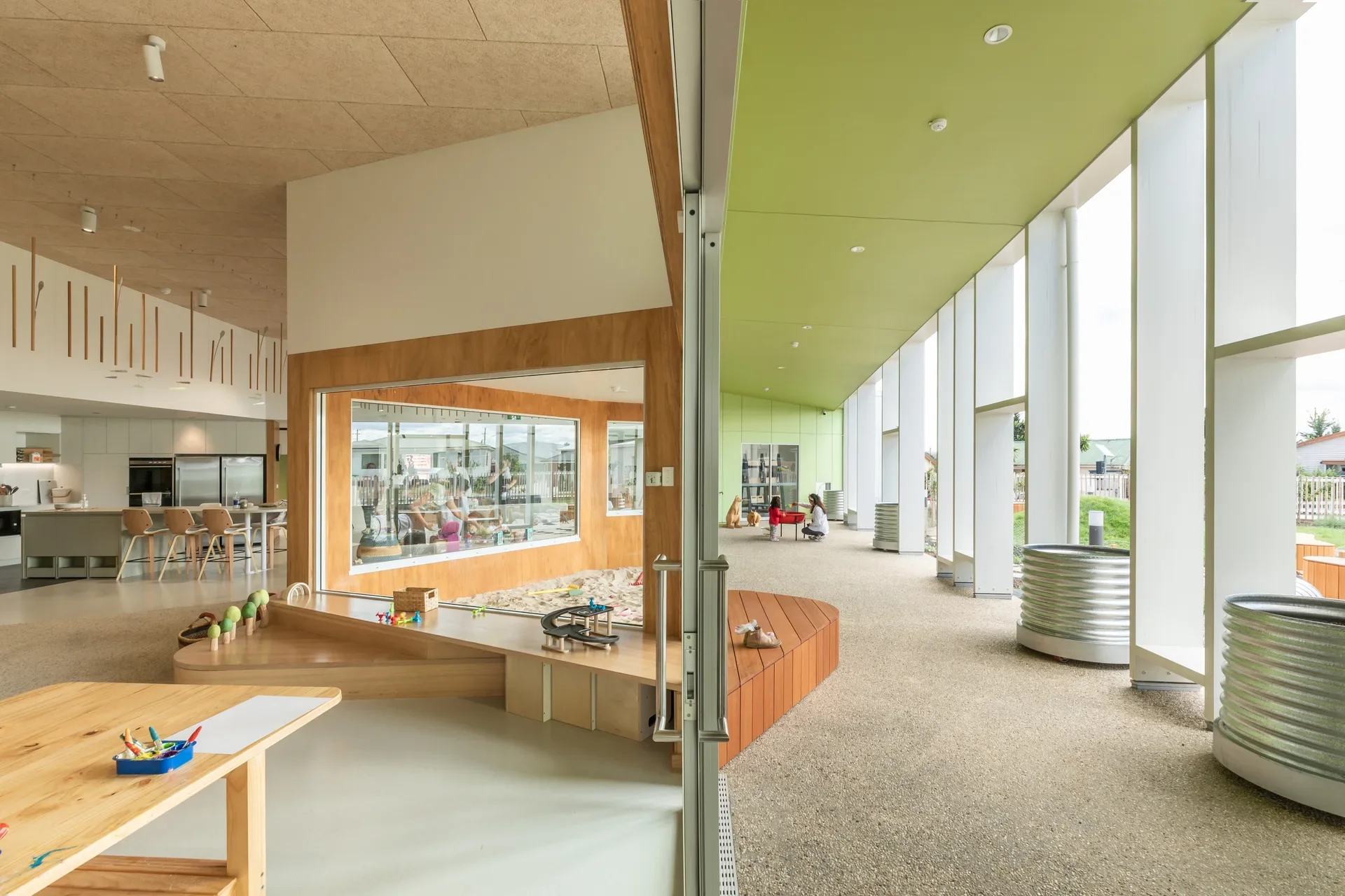 A vibrant, modern early learning centre featuring an indoor-outdoor flow. The space includes a lime-green ceiling, light timber play areas, and corrugated metal planters, all framed by large glass doors that connect the bright interior to a covered walkway.