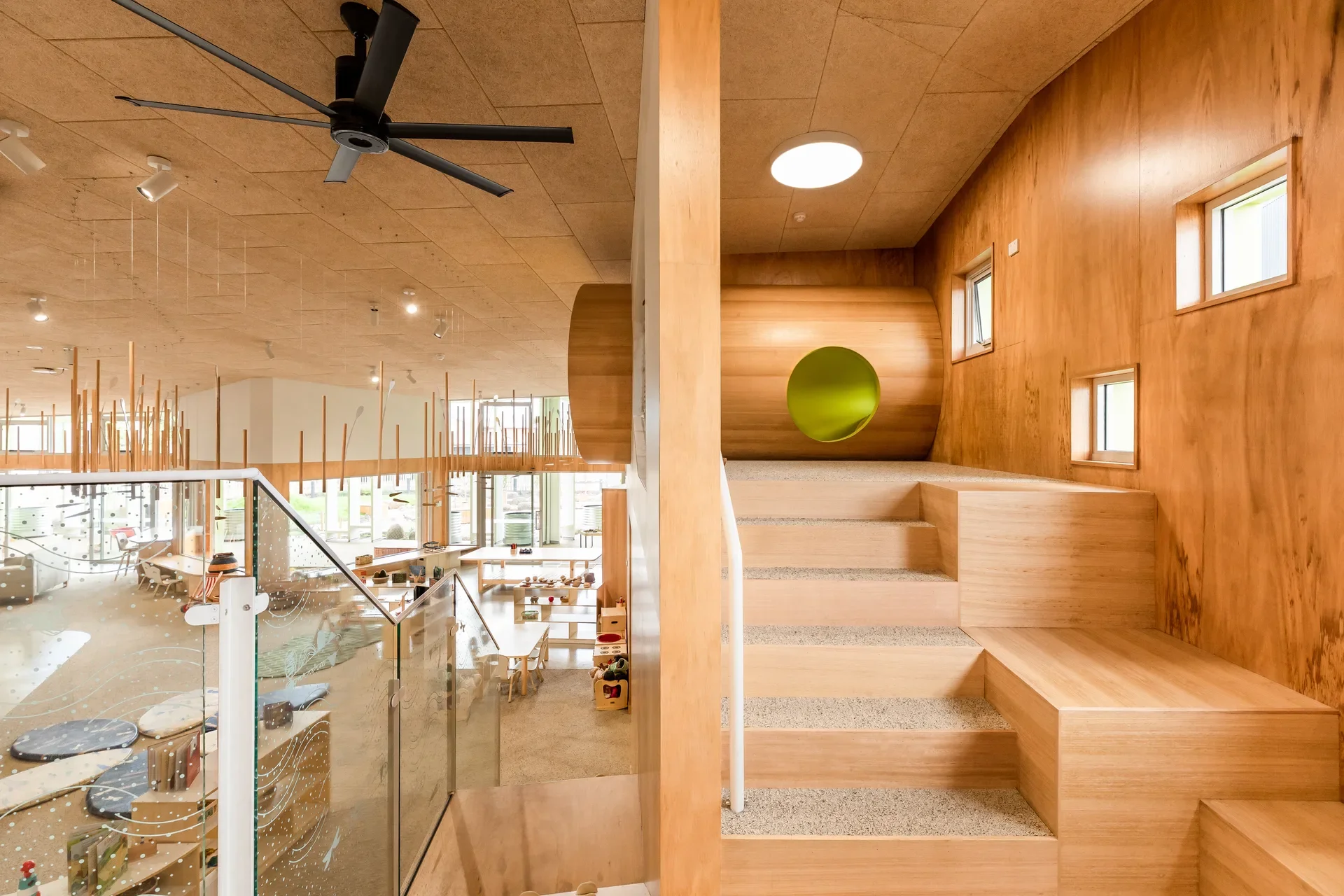 This interior features a wood-panelled landing with a staircase leading to a large wooden cylinder with a lime-green circular opening. A glass railing overlooks a lower level with decorative hanging rods and a black ceiling fan.