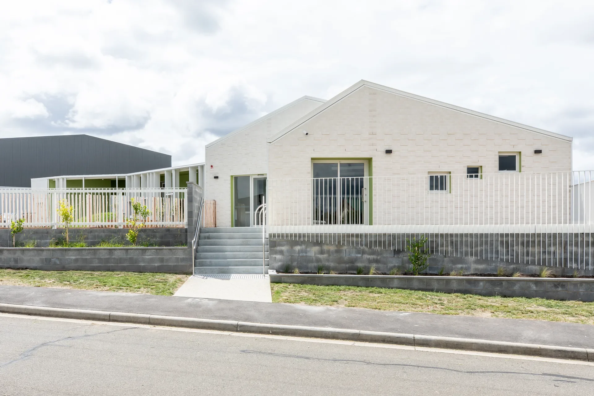 This modern facility features a textured white brick facade with a gabled roofline and green-trimmed windows. A set of grey concrete stairs leads from the sidewalk to a glass entrance, bordered by a white safety fence and a low dark-grey stone wall.