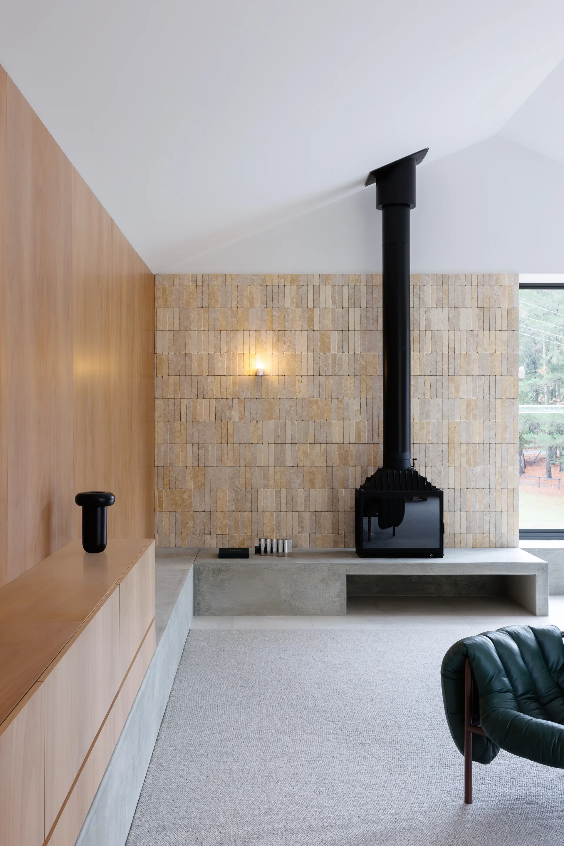 This modern living area features a black fire place with a tall flue, set against a wall of slender, vertical tan bricks. The fireplace sits on a low, concrete bench that extends along the base of a light wood-panelled wall, while a dark green tufted chair is partially visible in the foreground.