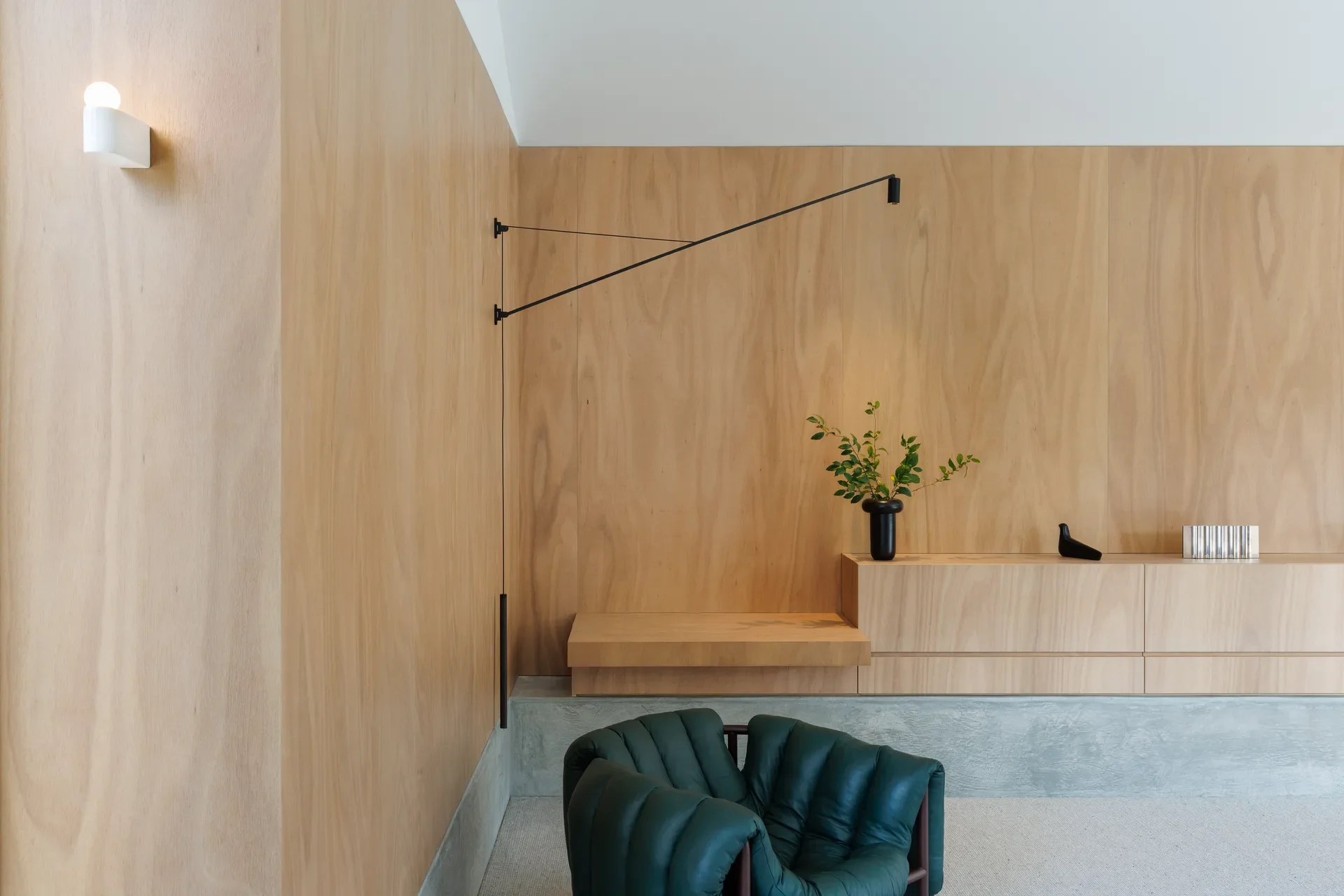 A minimalist interior featuring light-toned wood panelled walls and matching built-in cabinetry. A dark green, tufted leather armchair sits in the foreground on a neutral carpet. The space is accented by a slender black swing-arm wall lamp and a small black vase with green branches.